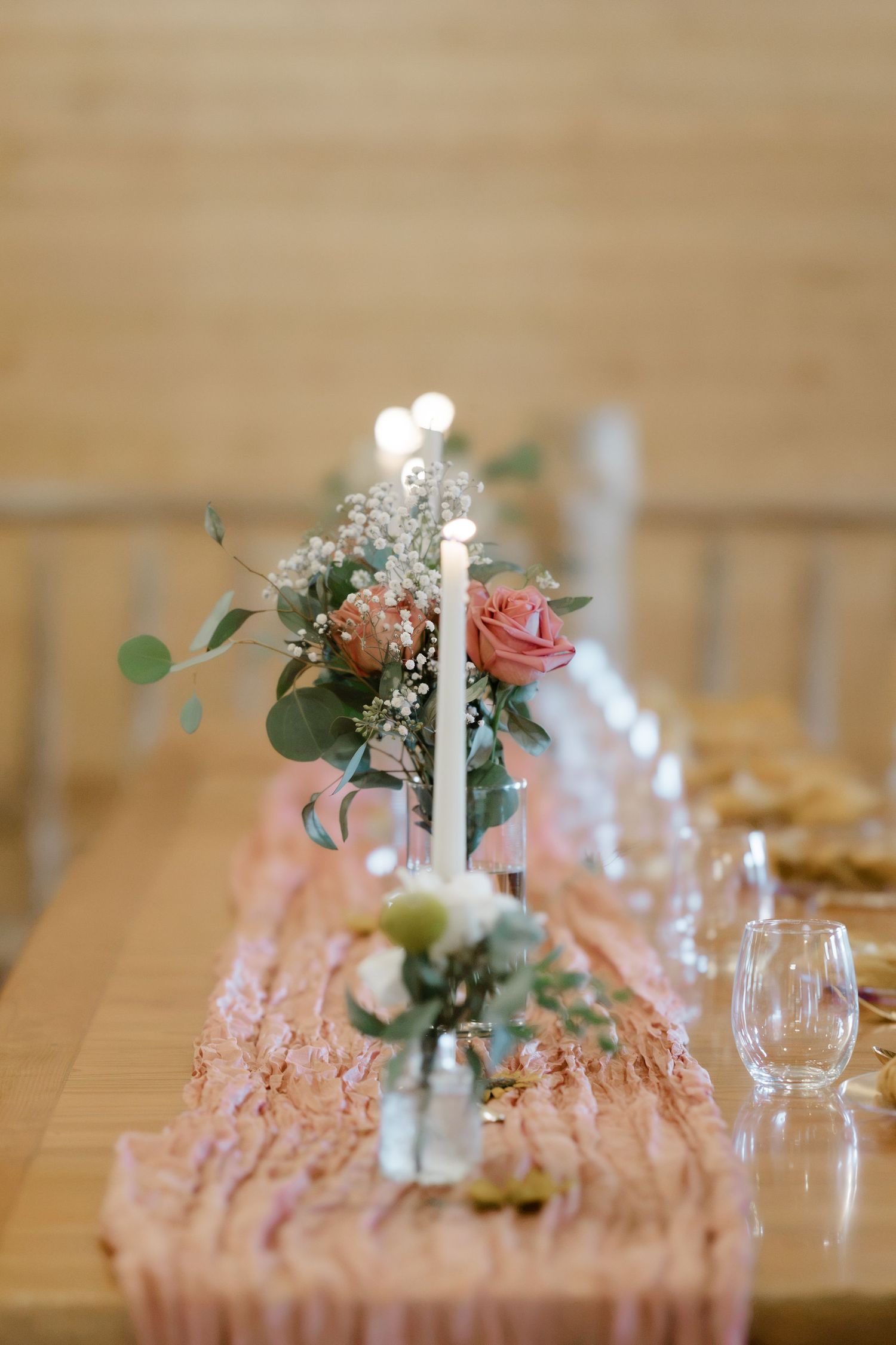 Elegant wedding table centerpiece with pink roses, baby's breath, eucalyptus, and a lit taper candle on a blush runner.