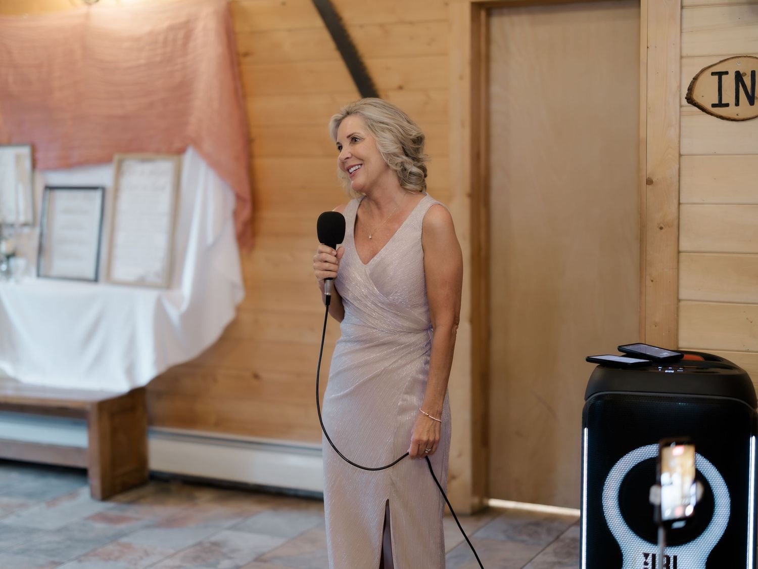 Elegant blonde woman in a champagne gown speaking into a microphone at an indoor wedding reception venue.