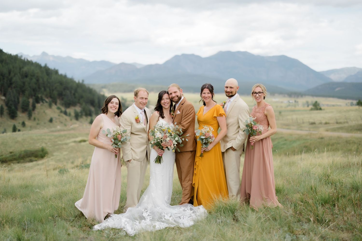 Wedding party poses in a mountain meadow, bride in white, bridesmaids in blush and gold, groomsmen in neutral tones.