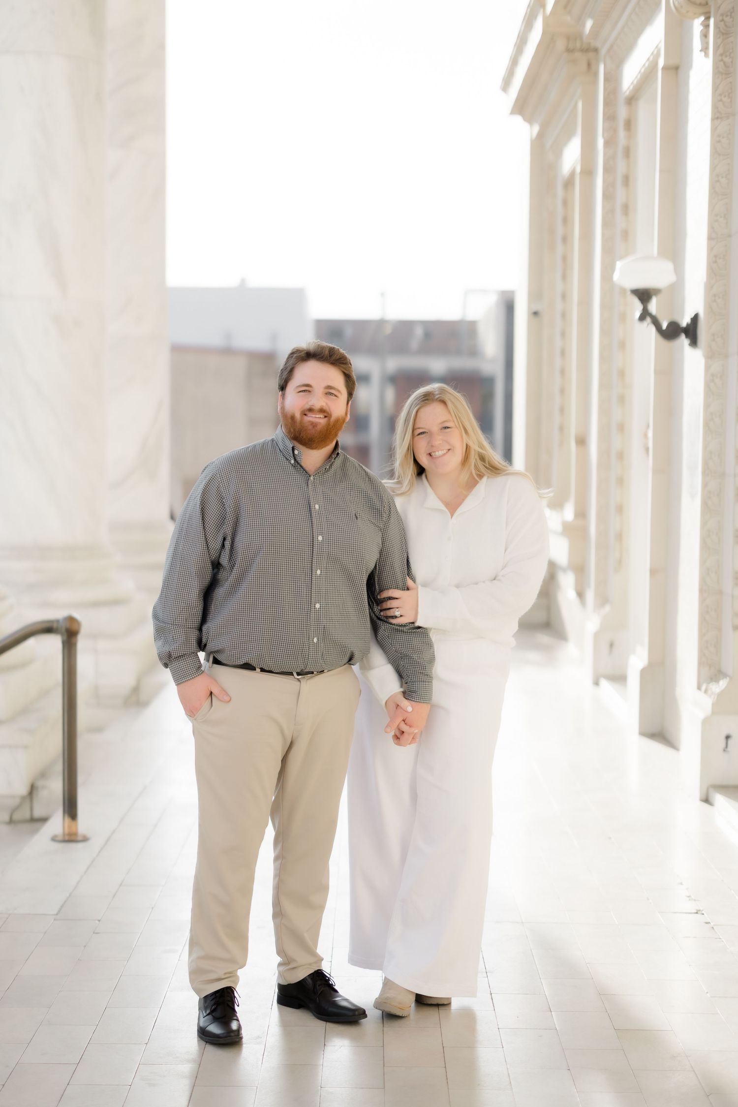Engaged couple holding hands and smiling while posing in a sunlit marble colonnade.