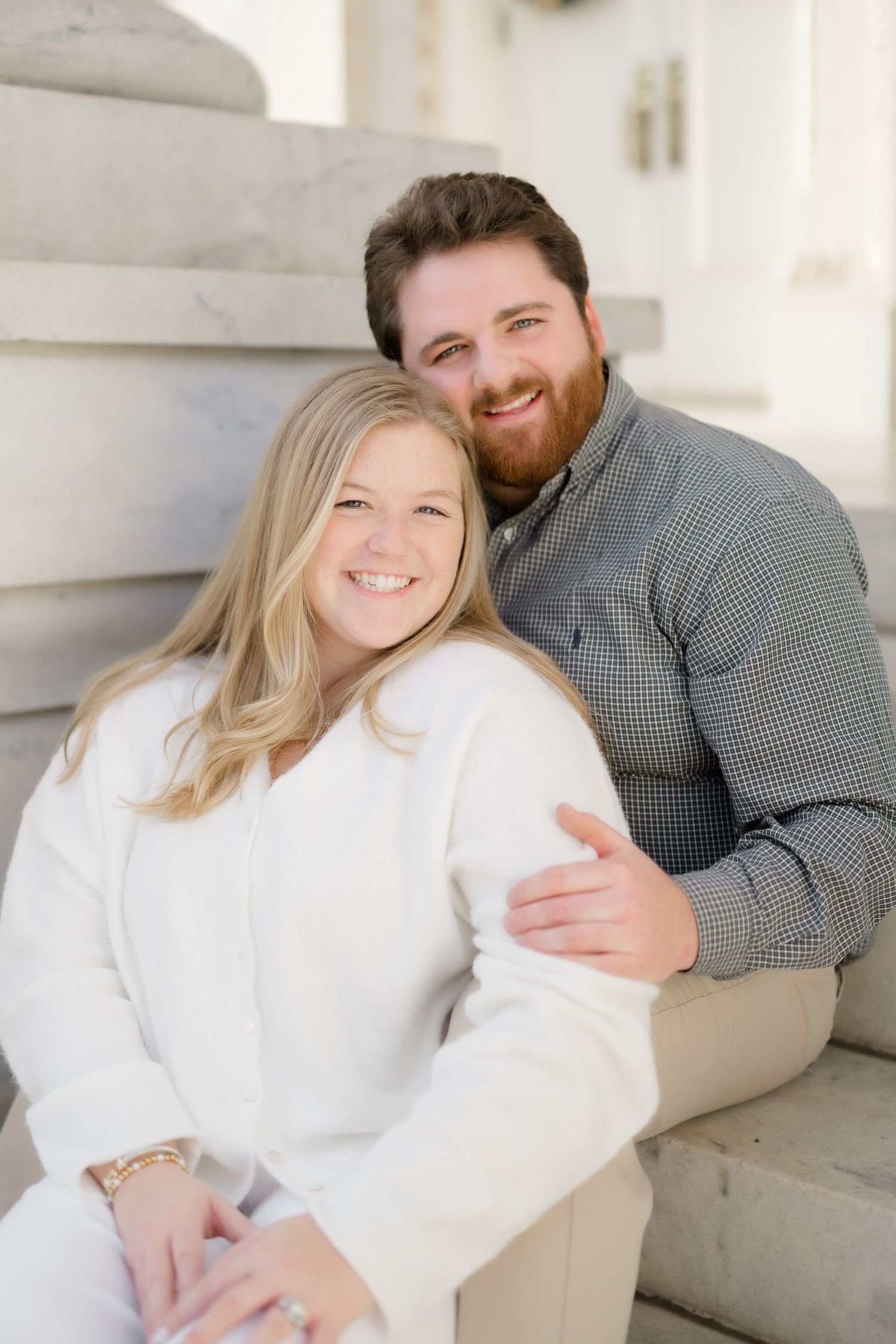 Smiling couple sitting on marble steps, woman in white outfit leaning against bearded man in gray shirt.