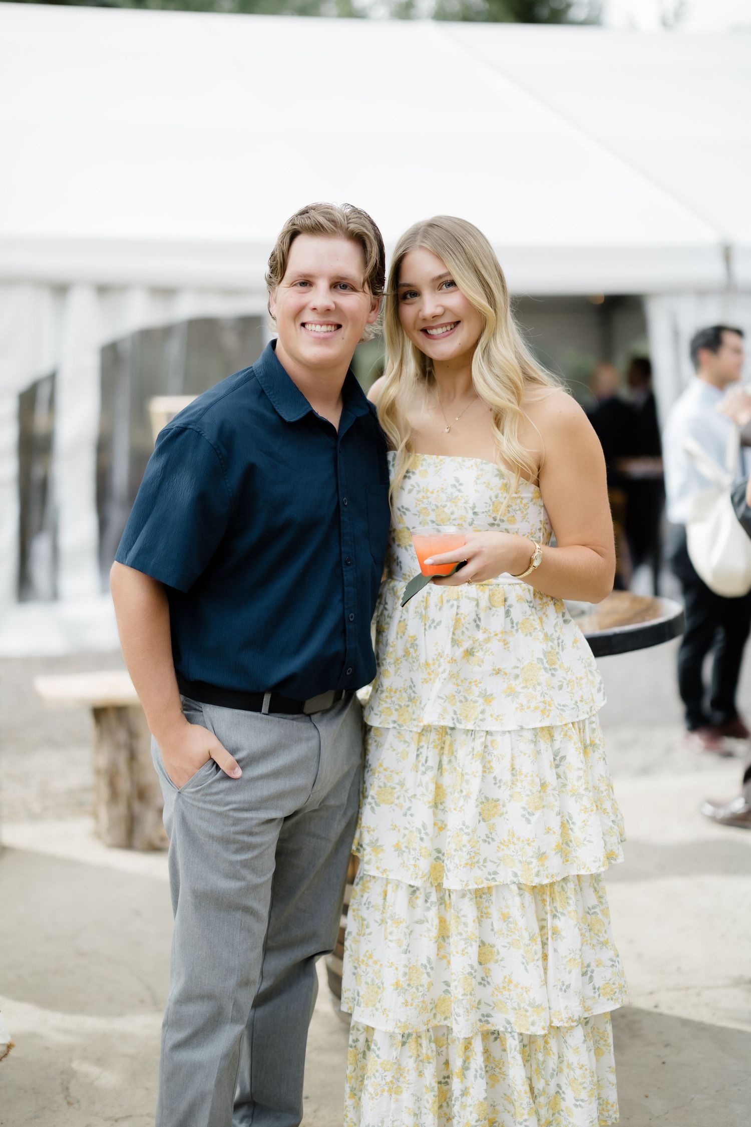 A smiling couple poses outdoors at an event, man in navy shirt and woman in floral tiered maxi dress.