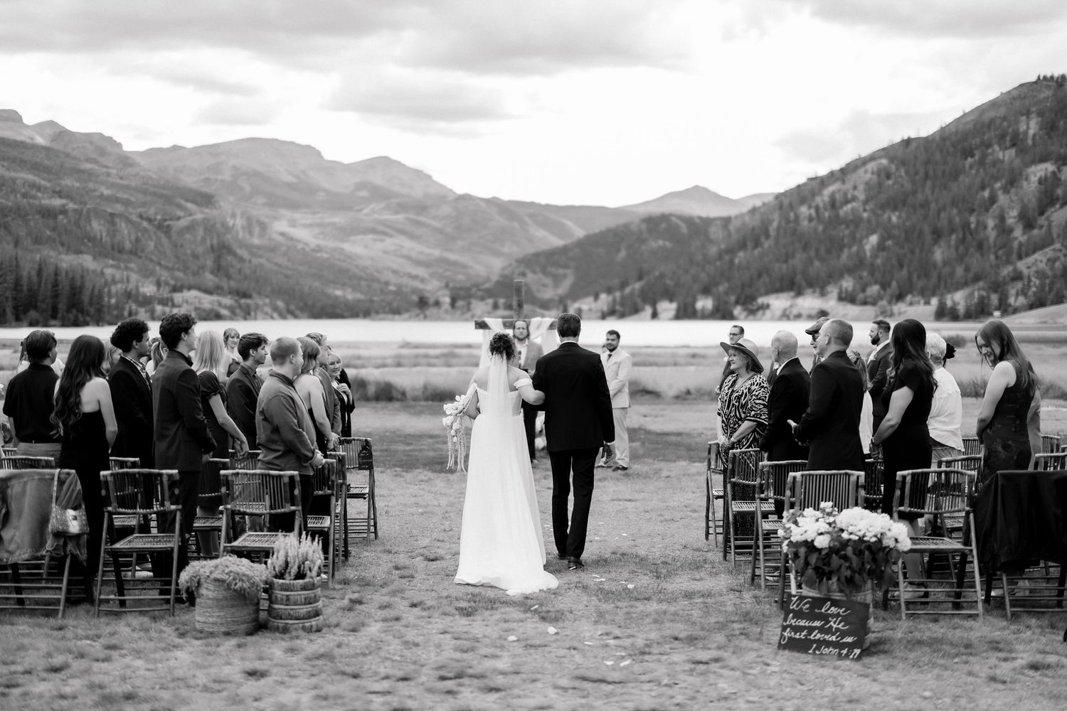 Black and white outdoor wedding ceremony in mountain valley with bride, groom, and seated guests.