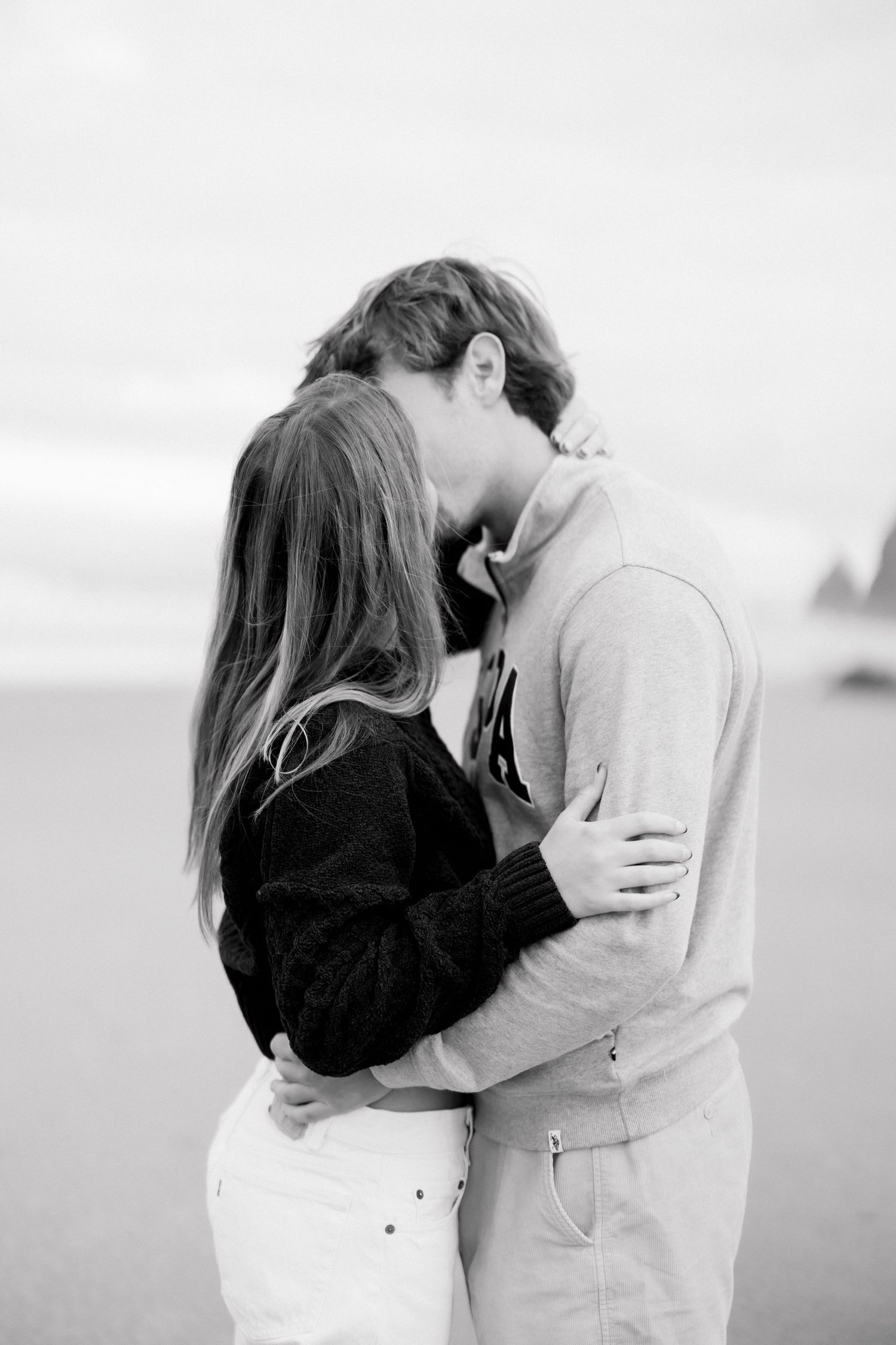 Black and white photo of a couple embracing and kissing on a beach, wearing casual sweaters and white pants.