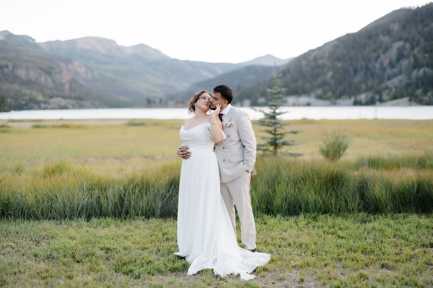Bride and groom embrace near a mountain lake meadow, with scenic Colorado peaks in the background.