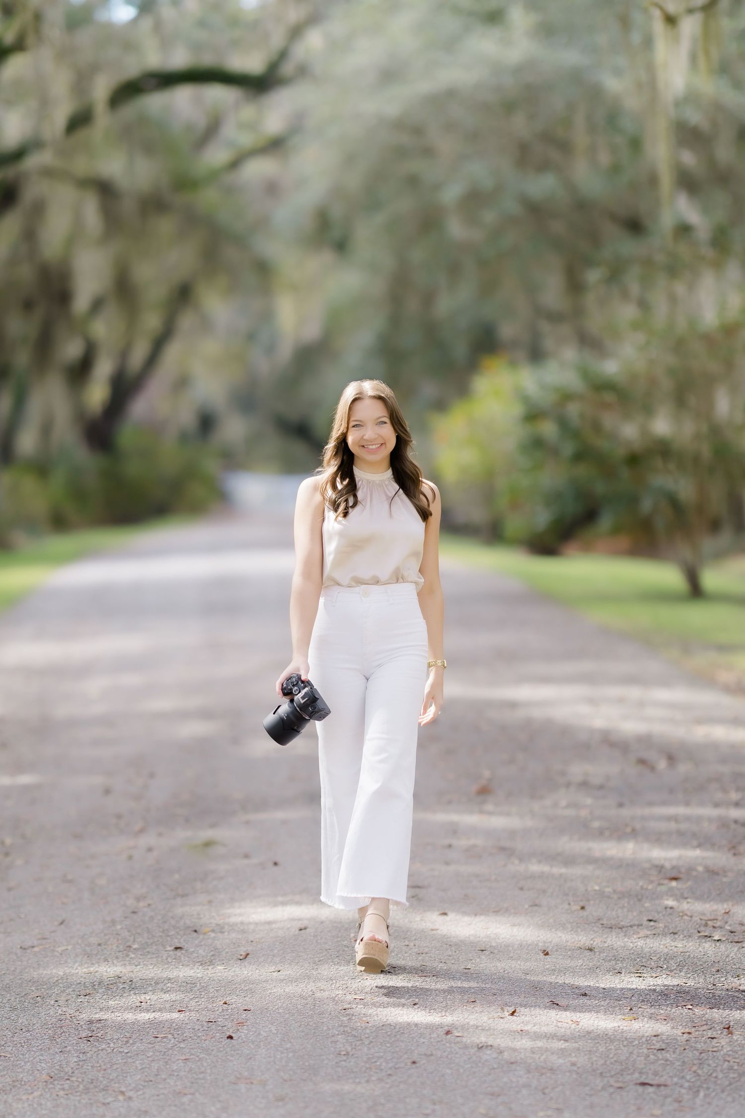Photographer in white dress walks along a tree-lined path holding a camera.