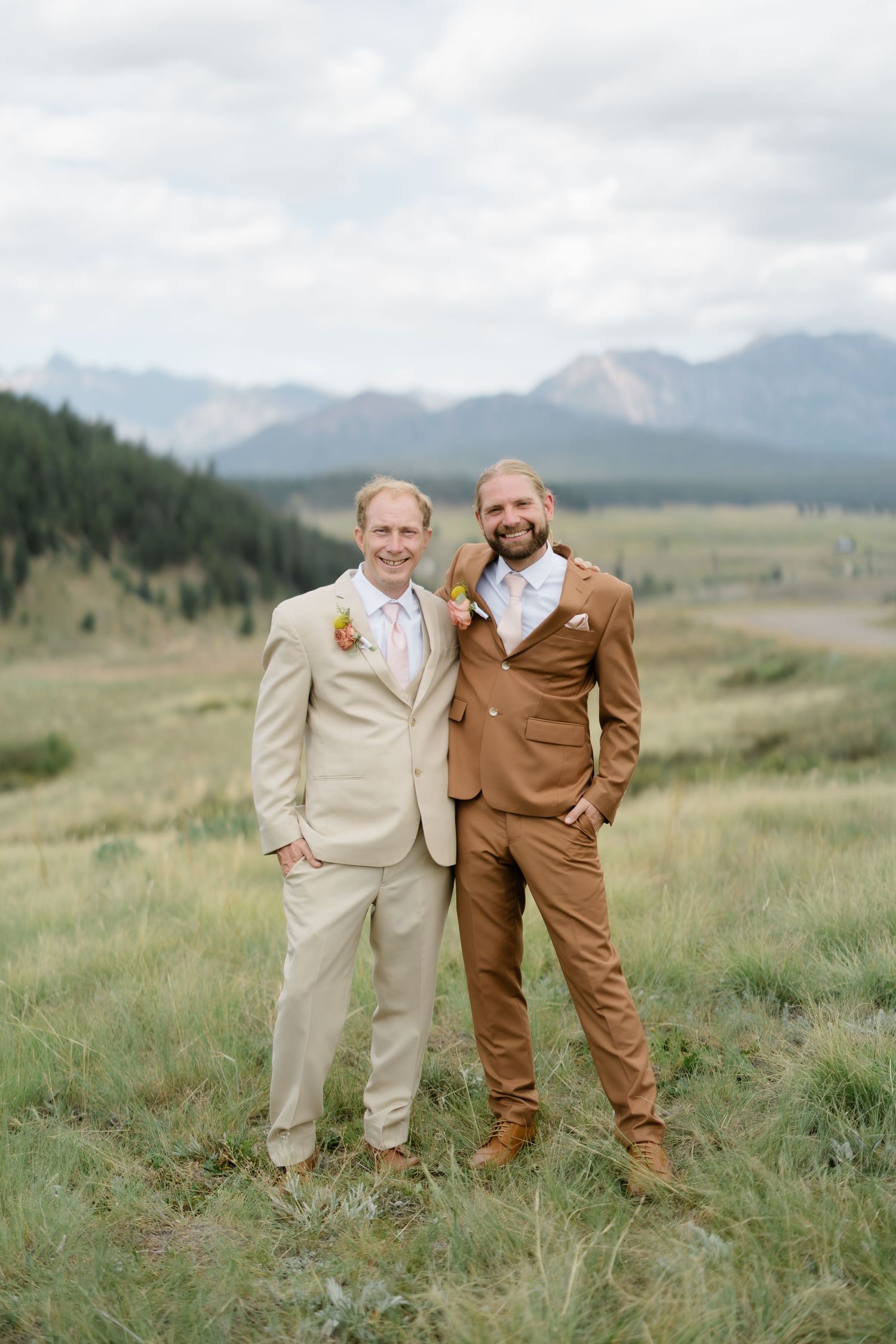Two grooms in tan and brown suits pose in a mountain meadow with Rocky Mountain peaks in the background.