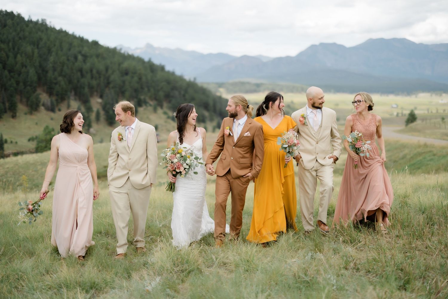Wedding party walks through mountain meadow, bride in white, bridesmaids in blush and gold, groomsmen in tan suits.
