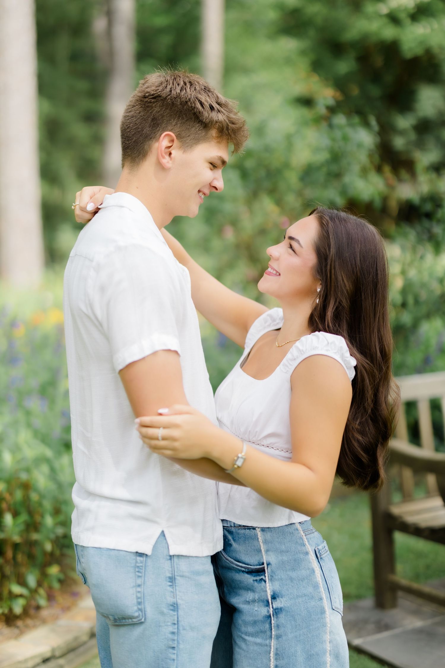A young couple in matching white tops and jeans embrace and smile at each other in a lush garden setting.