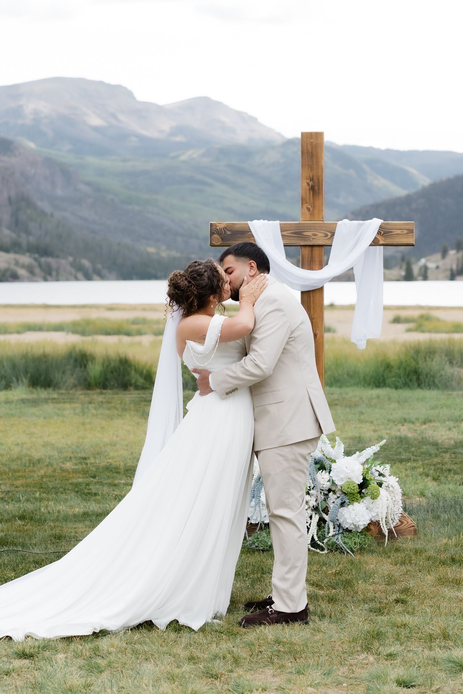 Bride and groom share first kiss at outdoor mountain wedding ceremony beside wooden cross with lake view.