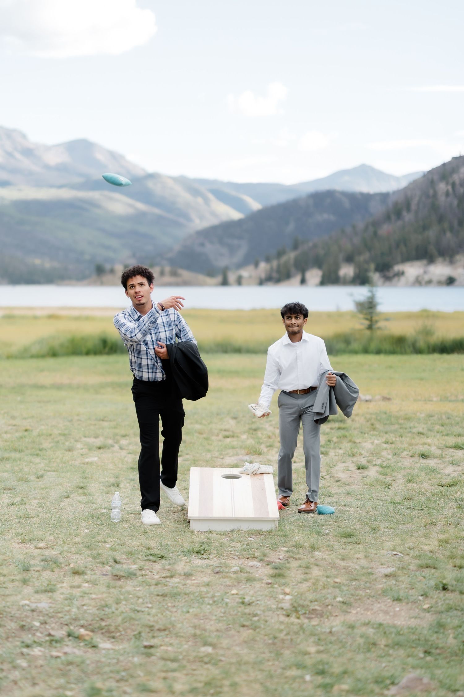 Two men playing cornhole outdoors near a mountain lake, one tossing a bean bag while the other watches.