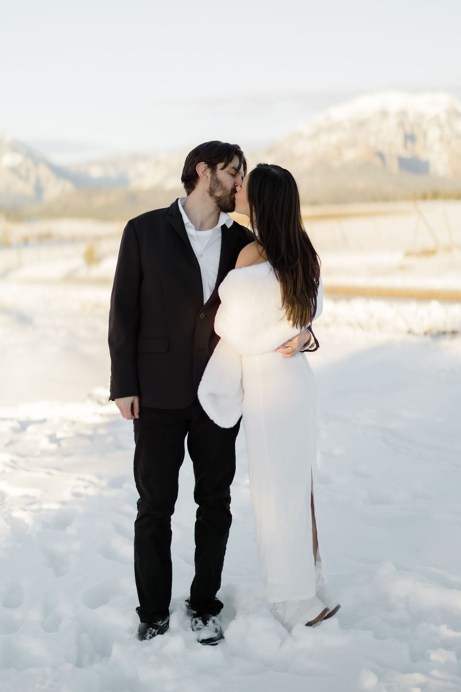 Bride and groom kissing in snowy mountain landscape, bride in white gown, groom in black suit.