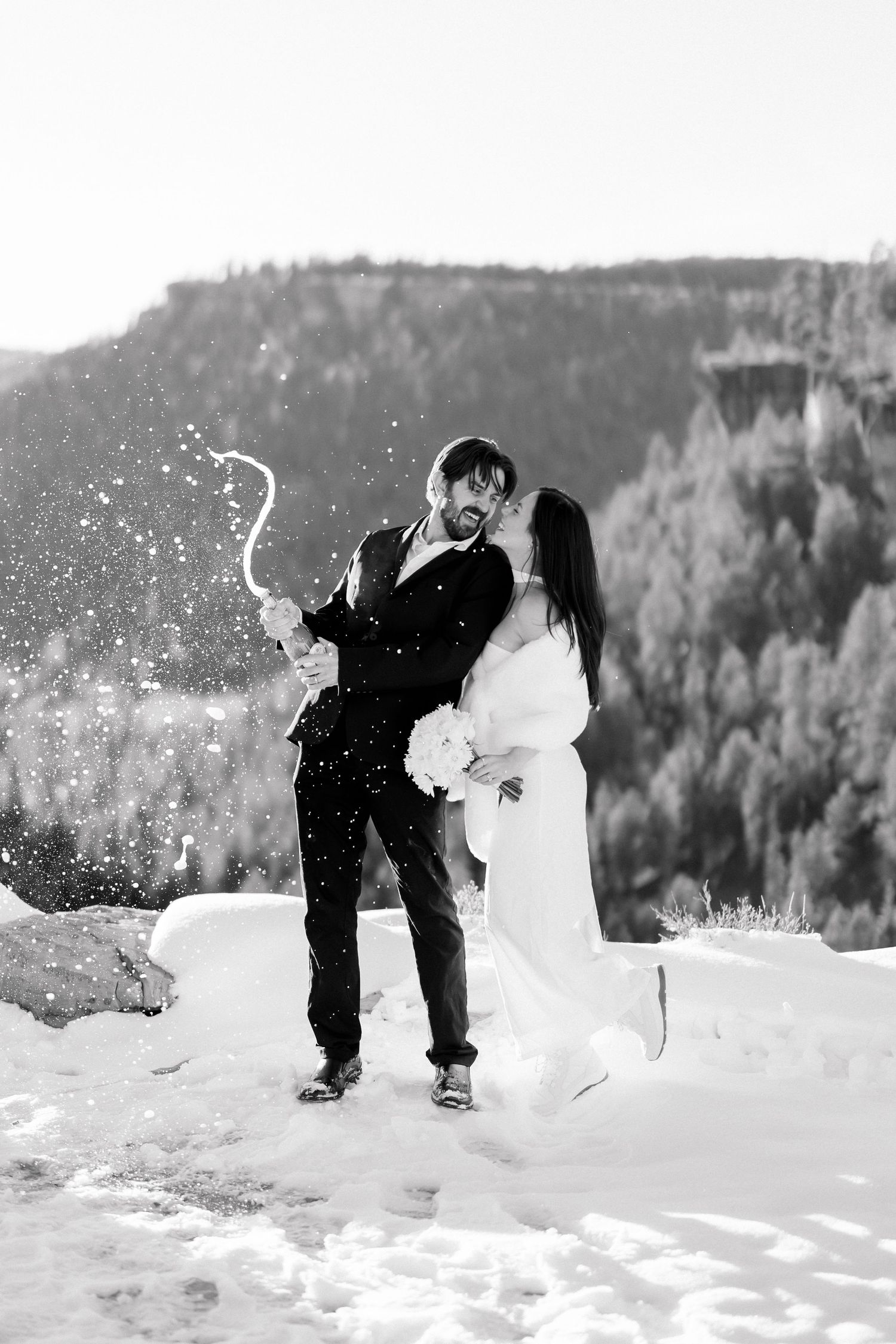 Black and white photo of a bride and groom popping champagne in the snow with a forested mountain backdrop.