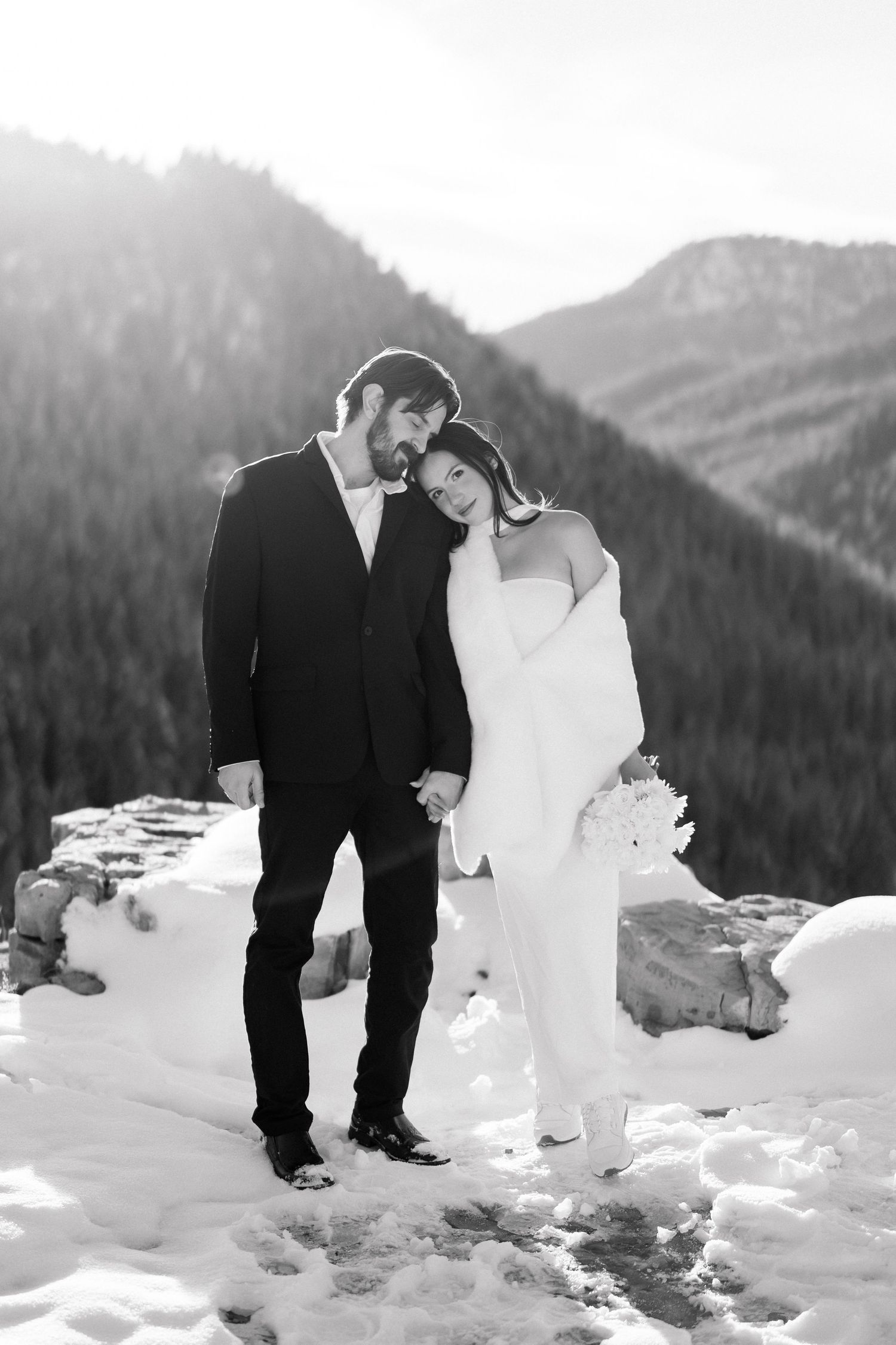 Black and white portrait of bride leaning on groom while holding bouquet in snowy mountain landscape.