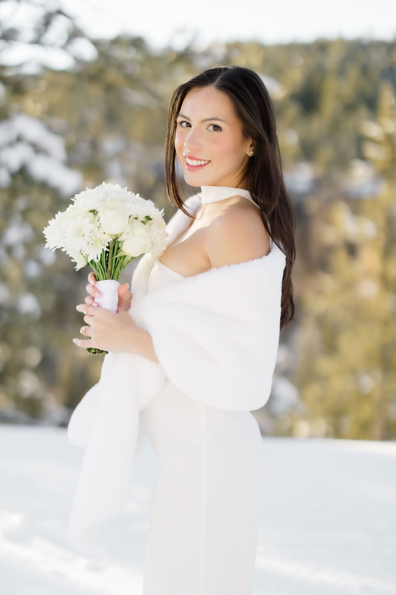 Radiant bride in white wedding gown and fur wrap smiling while holding white bouquet in winter snow.
