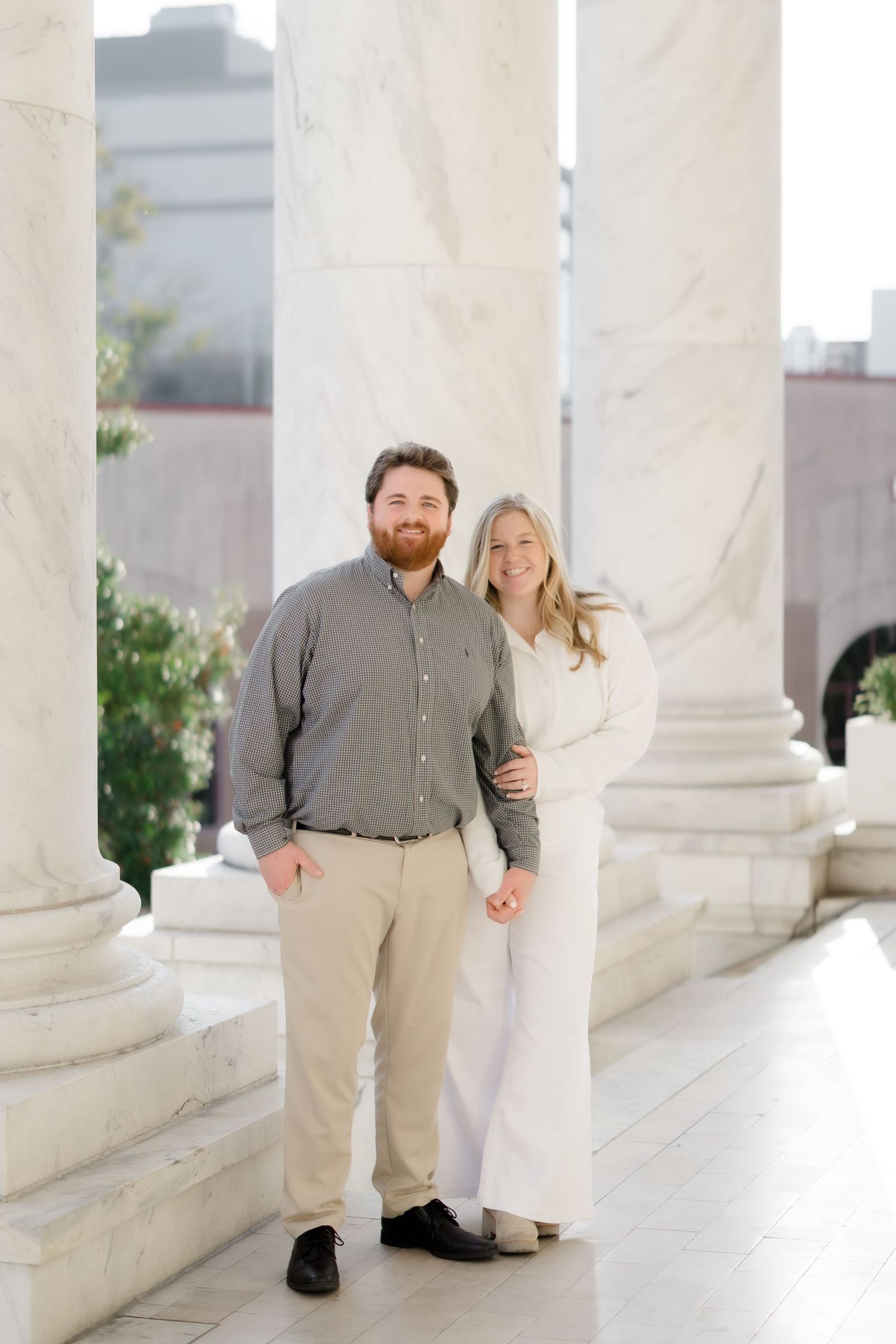 Couple posing between white marble columns, man in gray shirt and khakis, woman in white outfit, elegant outdoor setting.
