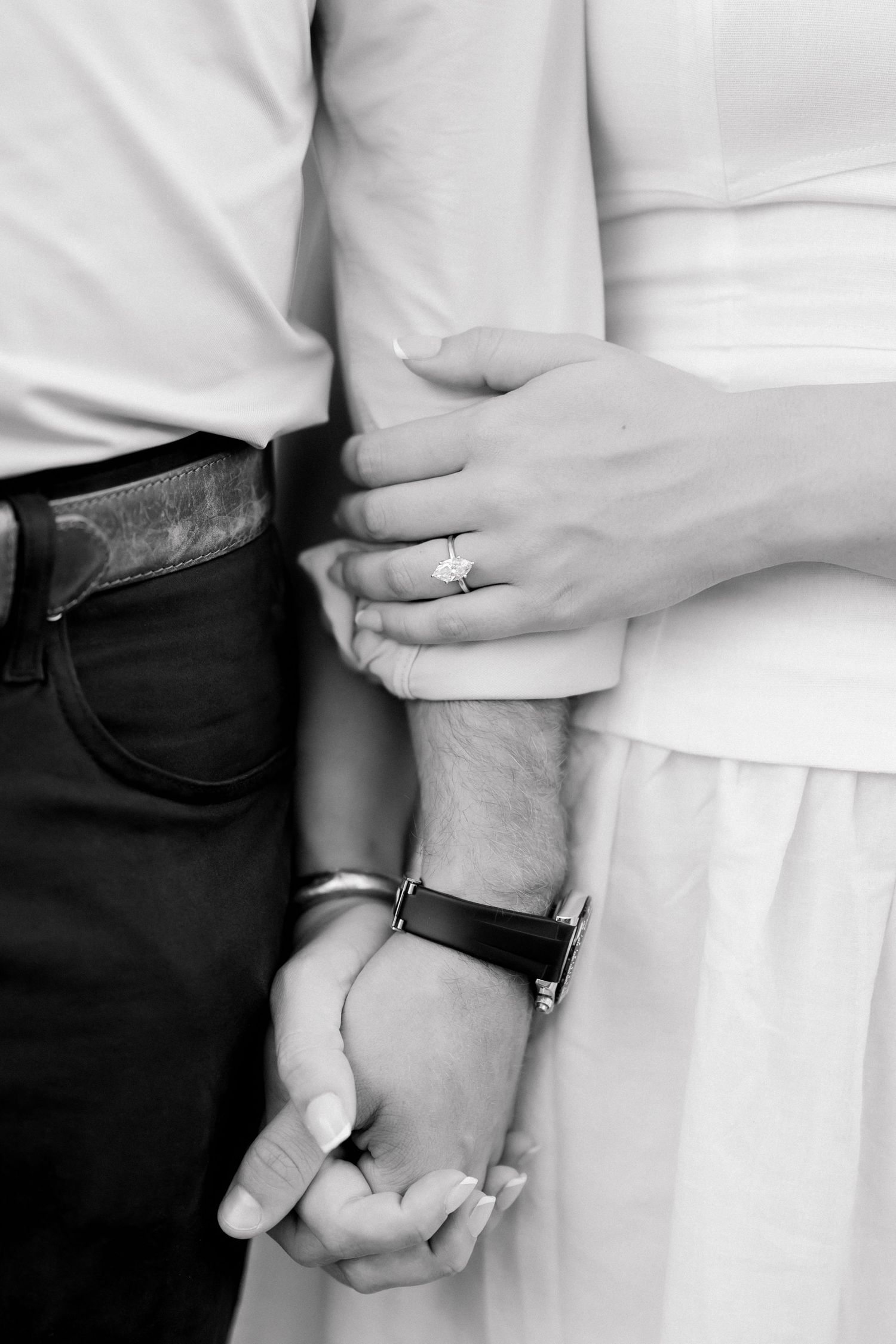 Black and white photo of couple holding hands, woman wearing diamond engagement ring, man wearing leather watch.