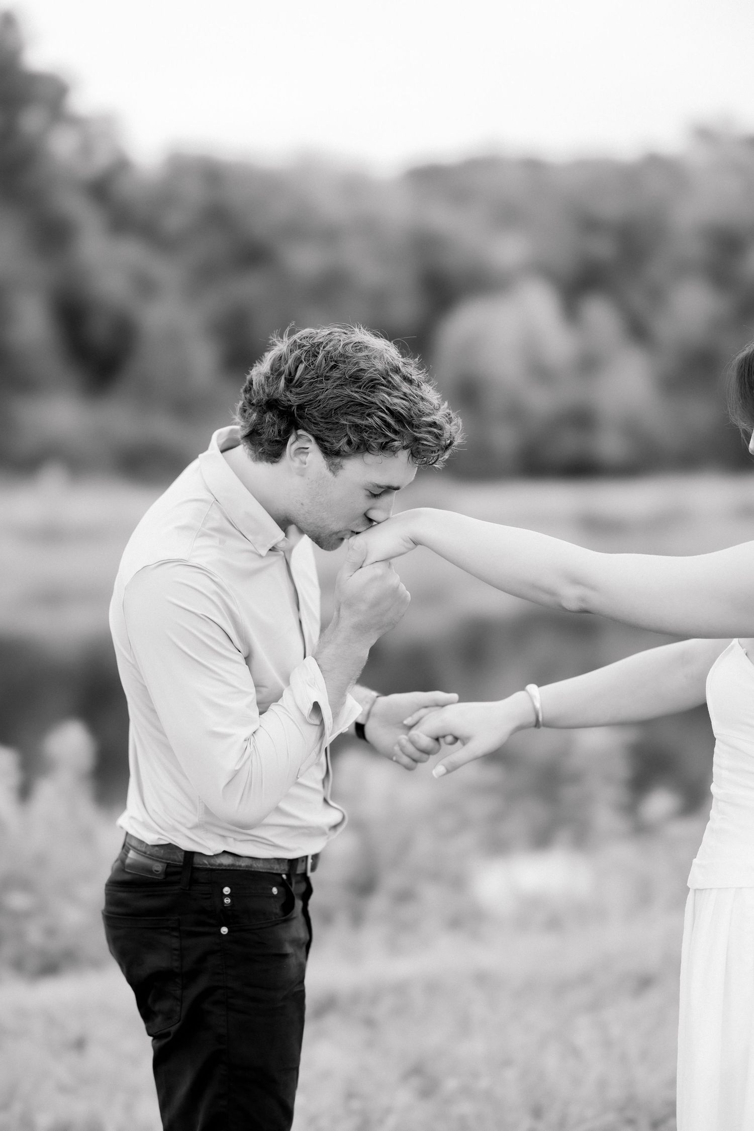 Black and white photo of a young man kissing a woman's hand outdoors near a lake.