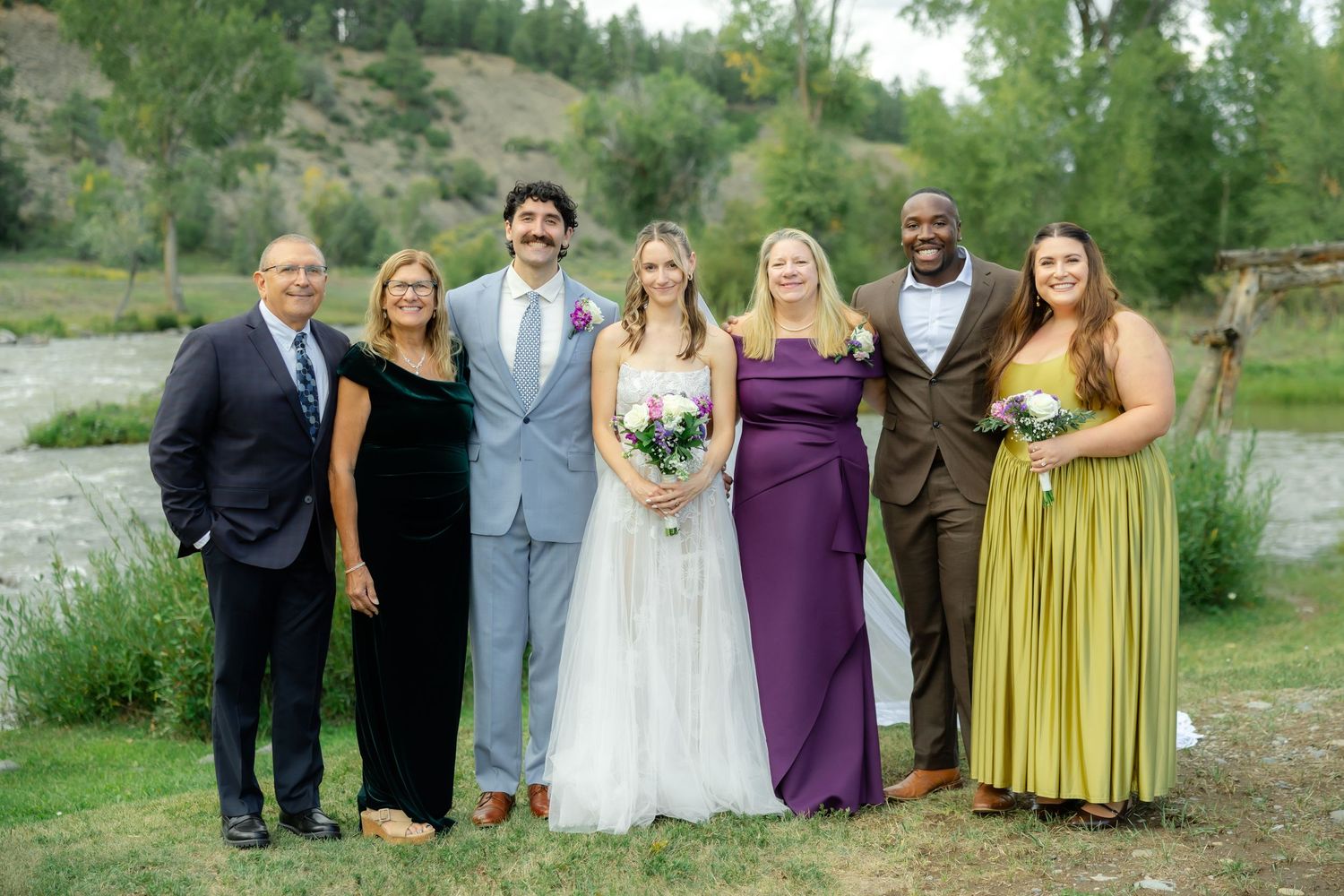 Wedding party poses outdoors near a river, featuring bride and groom with family in formal attire.