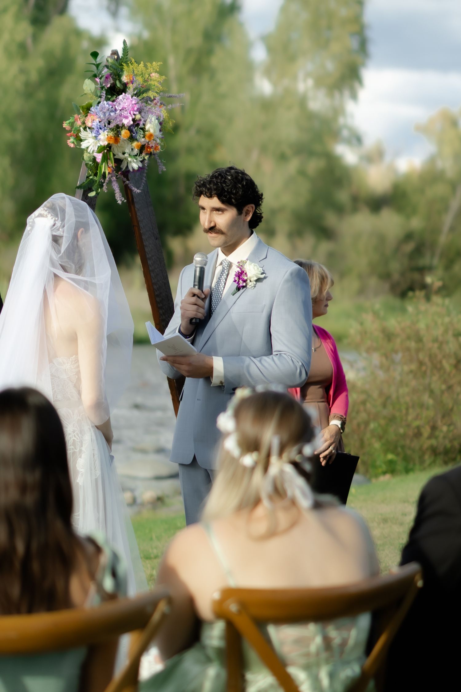 Groom in gray suit reads vows at outdoor wedding ceremony with floral arch, bride in white veil stands opposite.
