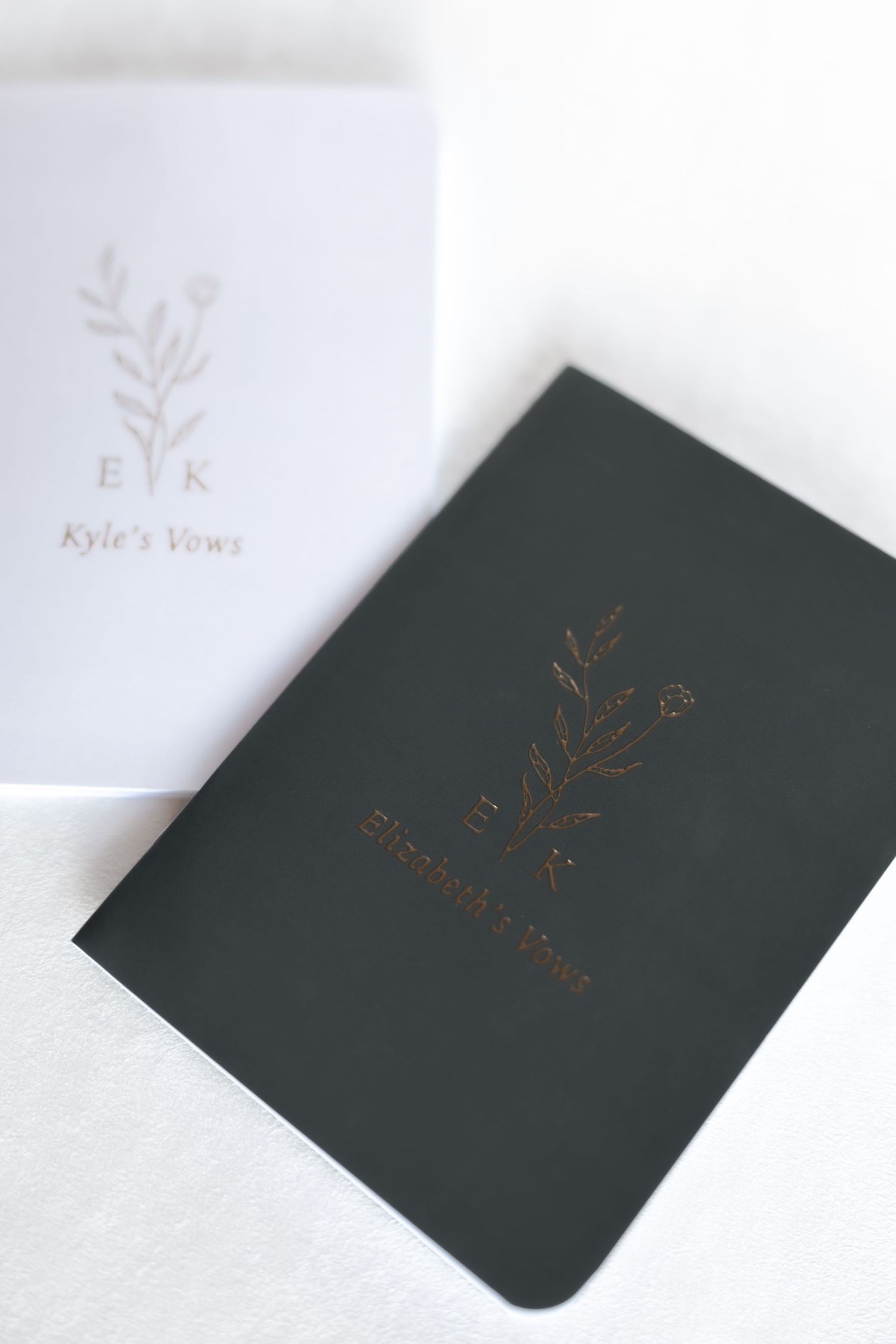 Two personalized wedding vow books in white and dark green with gold foil botanical monogram designs.
