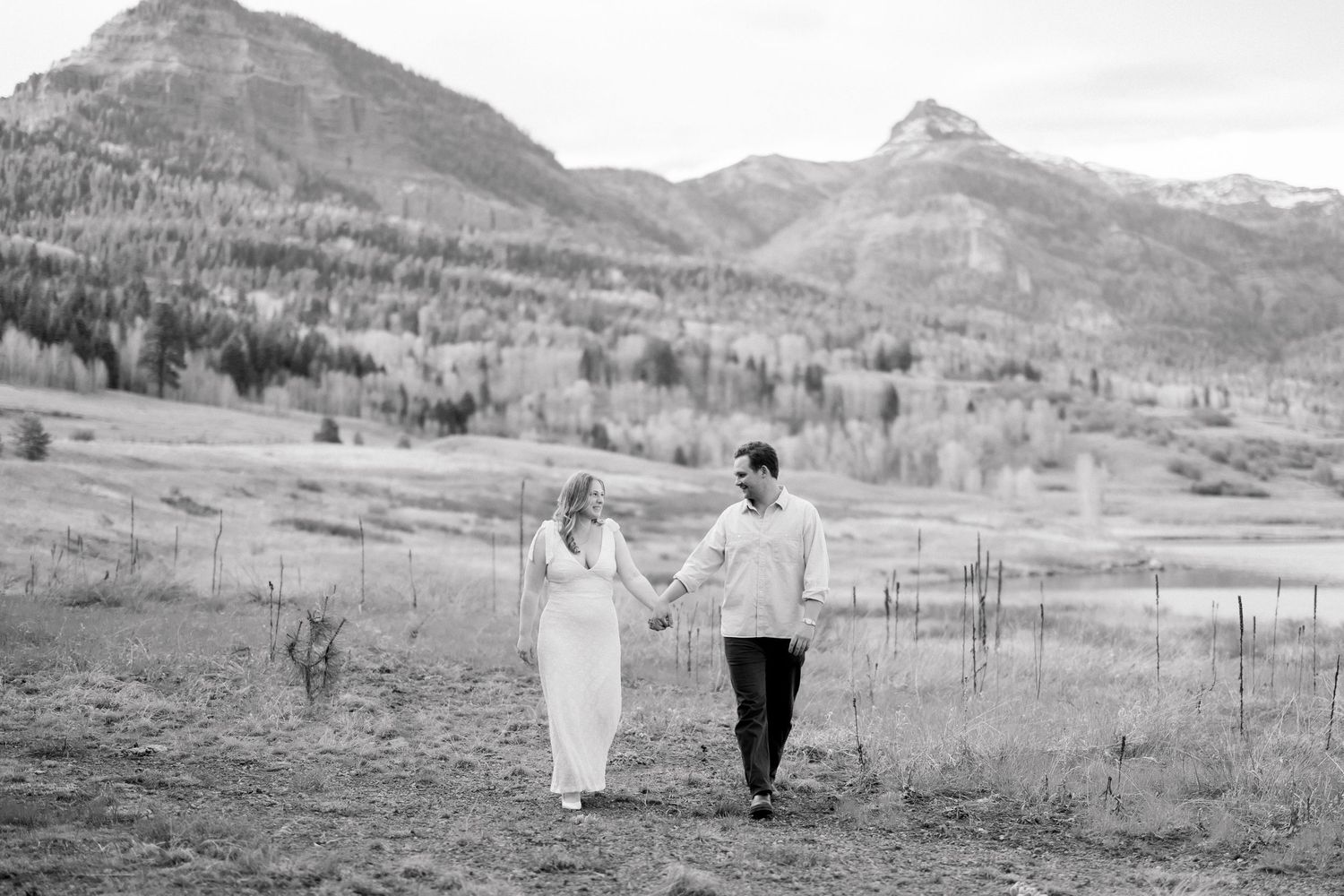 Black and white photo of a couple holding hands walking through a mountain meadow with scenic peaks in the background.