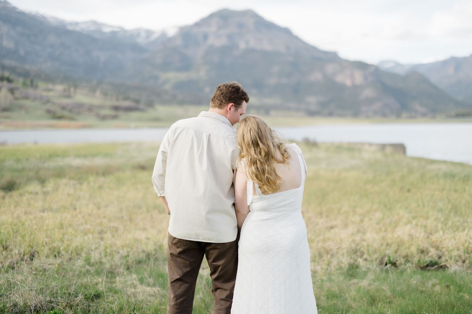 A couple stands together gazing at a mountain lake landscape, the woman in a white dress and man in a cream shirt.