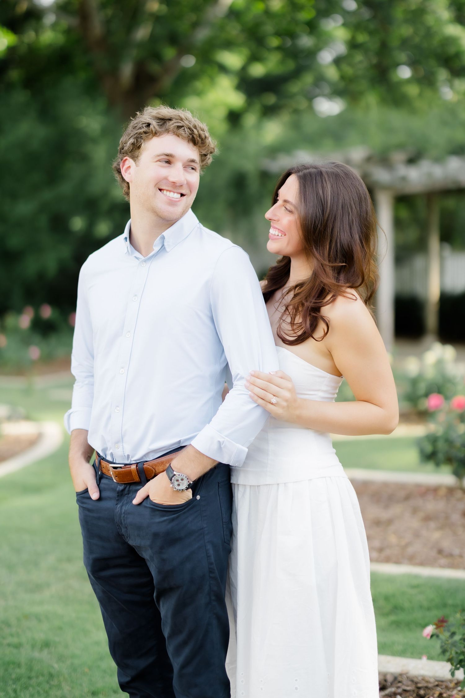 A smiling couple poses outdoors in a lush garden, woman in white dress embracing man in blue shirt and navy pants.