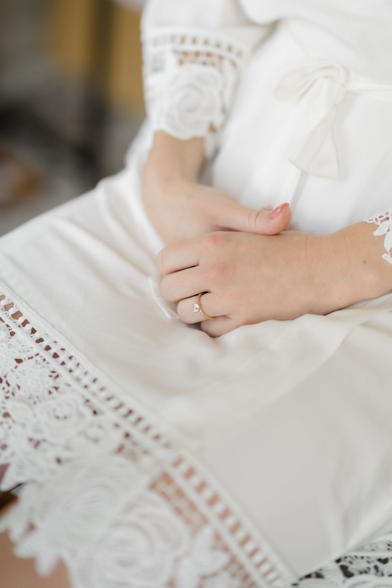 Close-up of a bride's hands folded in her lap wearing an engagement ring, dressed in a white lace wedding gown.