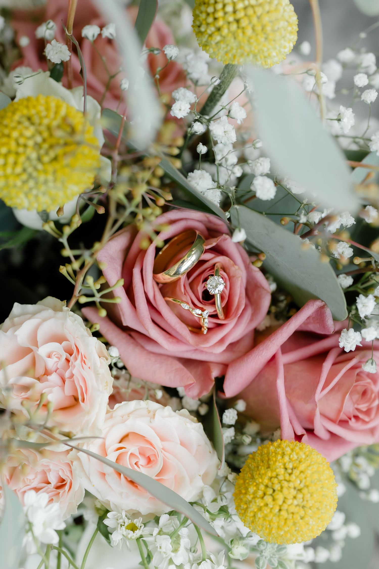 Gold wedding rings rest inside a pink rose in a bridal bouquet with yellow billy balls and baby's breath.
