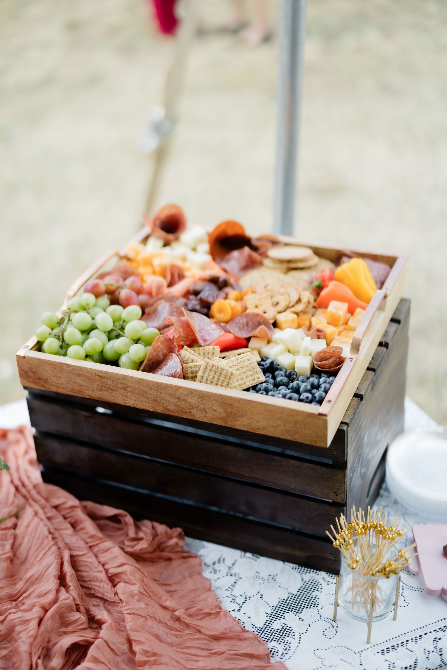 Rustic wooden crate filled with a charcuterie board featuring grapes, blueberries, meats, cheeses, and crackers outdoors.