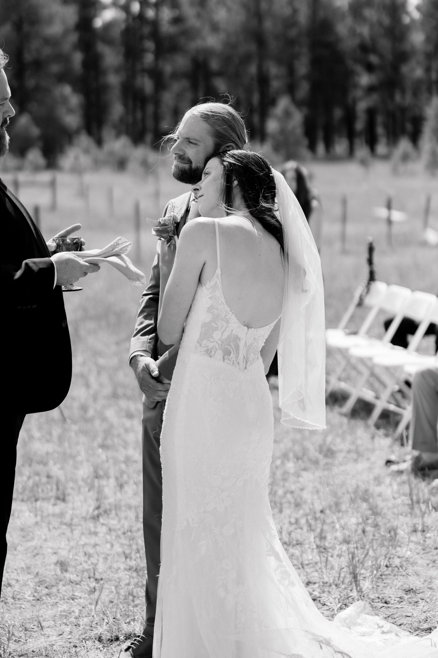 Black and white photo of a bride and groom standing at their outdoor wedding ceremony, bride in lace backless gown with veil.
