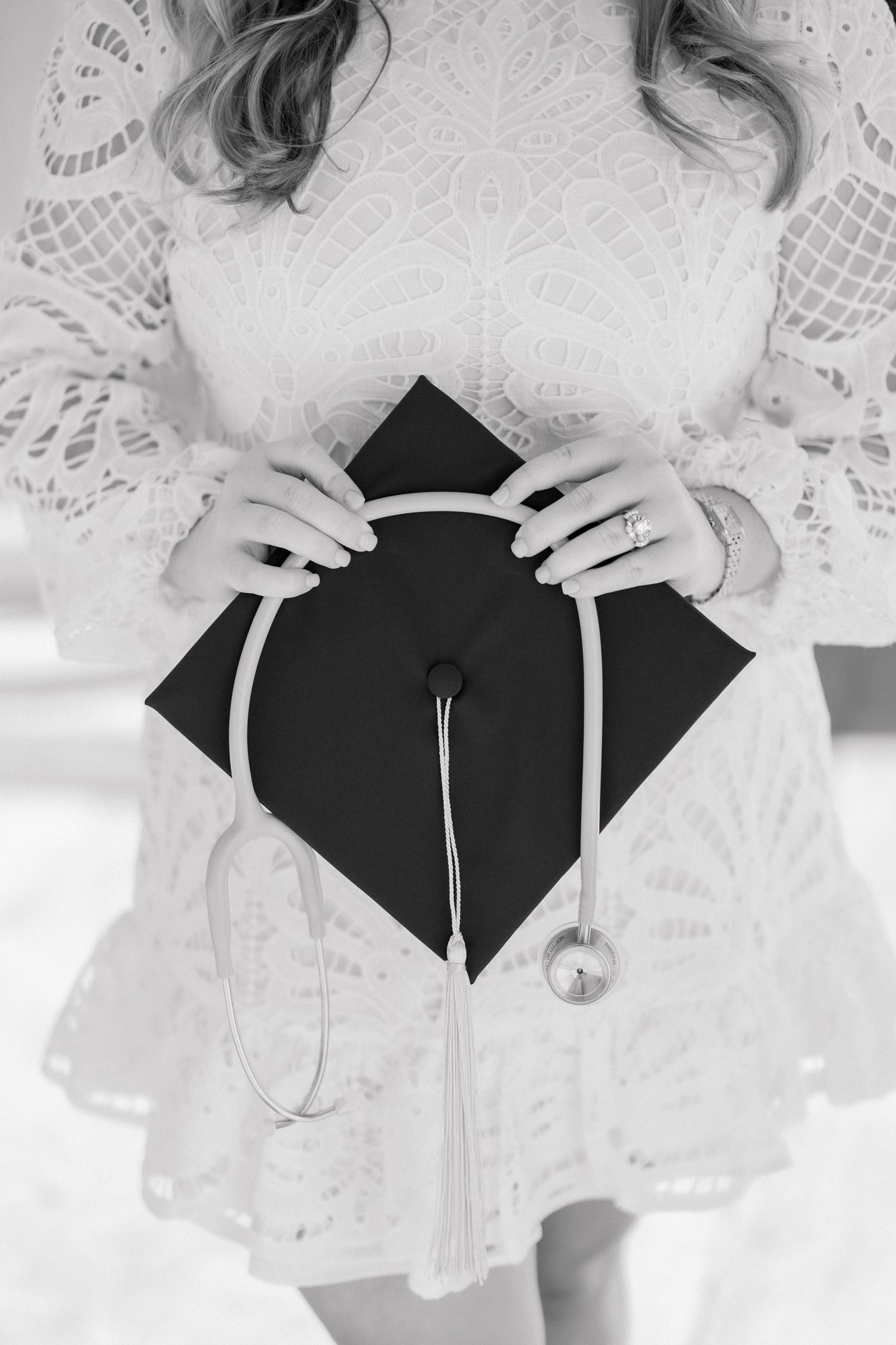 Black and white photo of a woman in white lace dress holding a graduation cap with stethoscope draped over it.