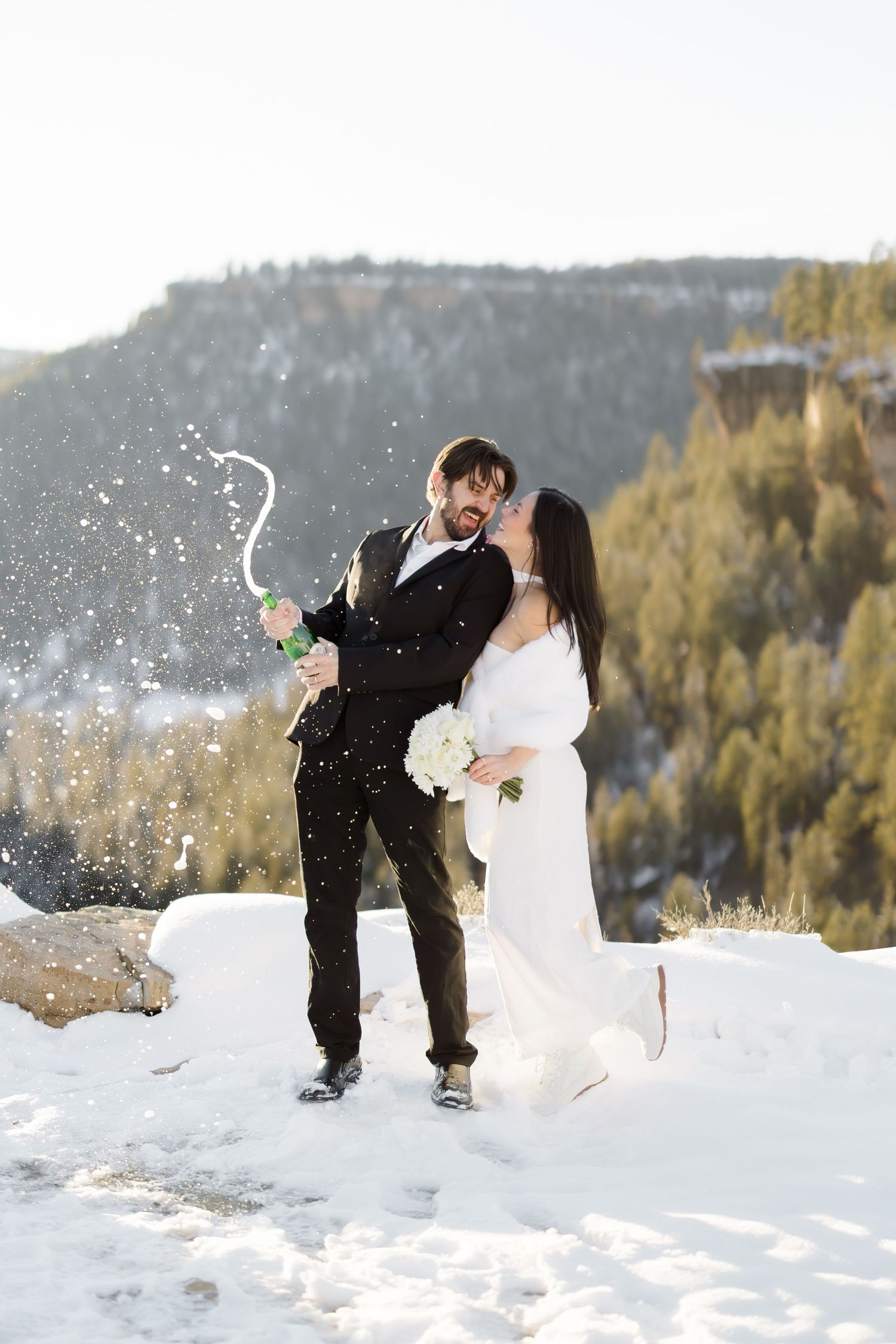 Newlyweds kissing while popping champagne bottle in snowy mountain landscape during winter wedding celebration.