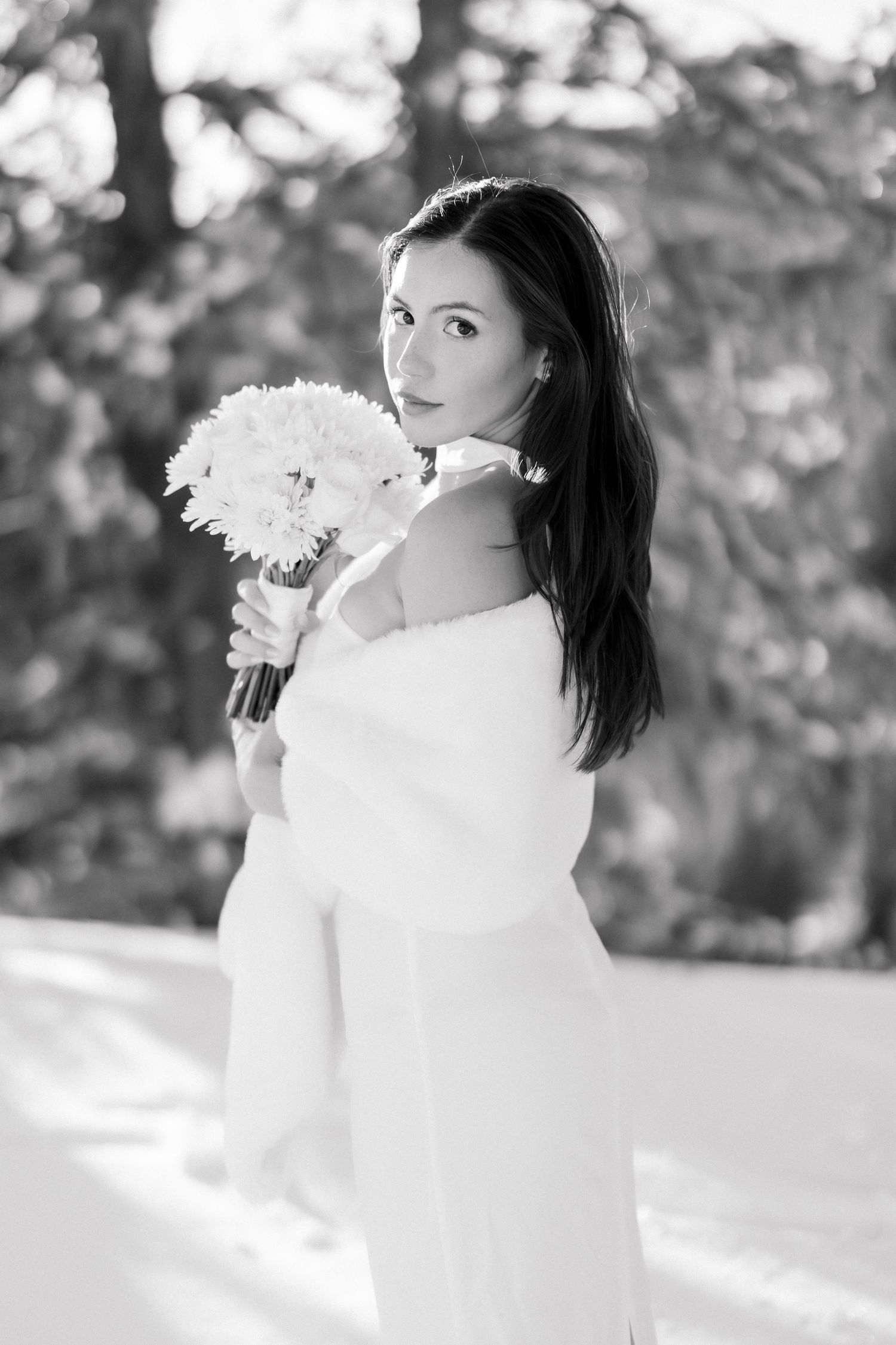 Black and white portrait of bride in off-shoulder fur wrap holding white bouquet in snow.