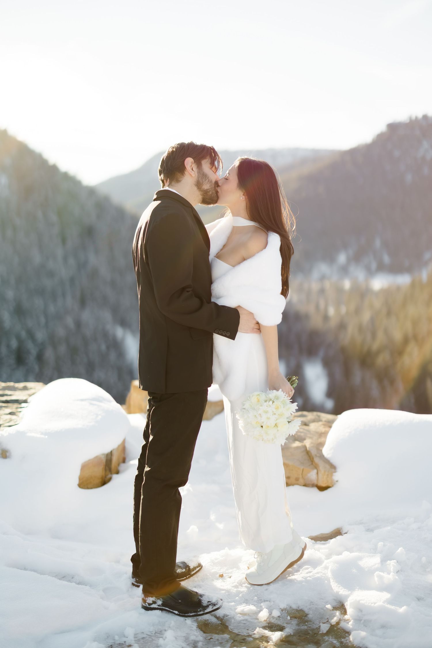 Bride and groom share a kiss in snowy mountain landscape, bride in white gown with fur wrap holding bouquet.