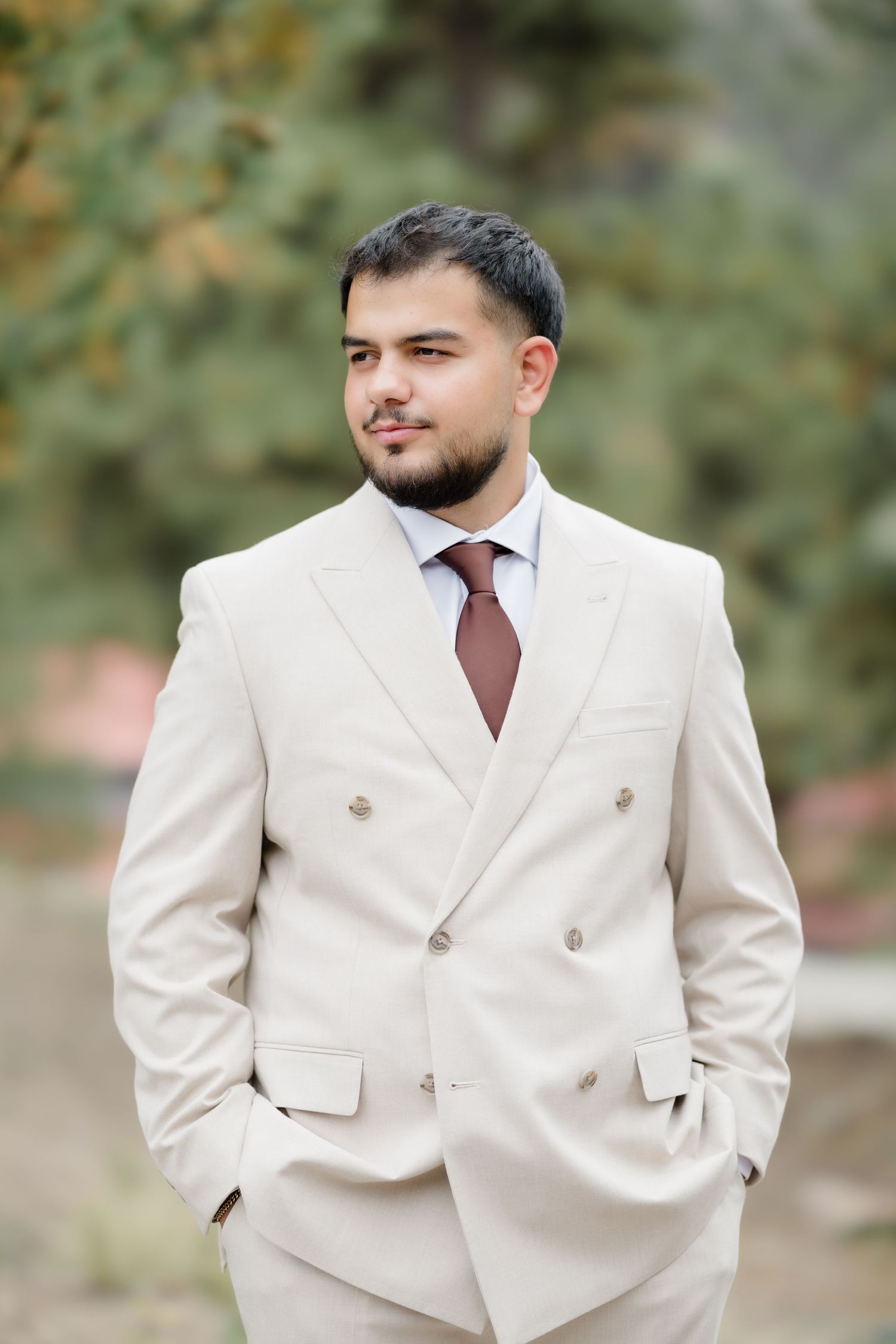 Young man in beige double-breasted suit with burgundy tie standing outdoors with hands in pockets.