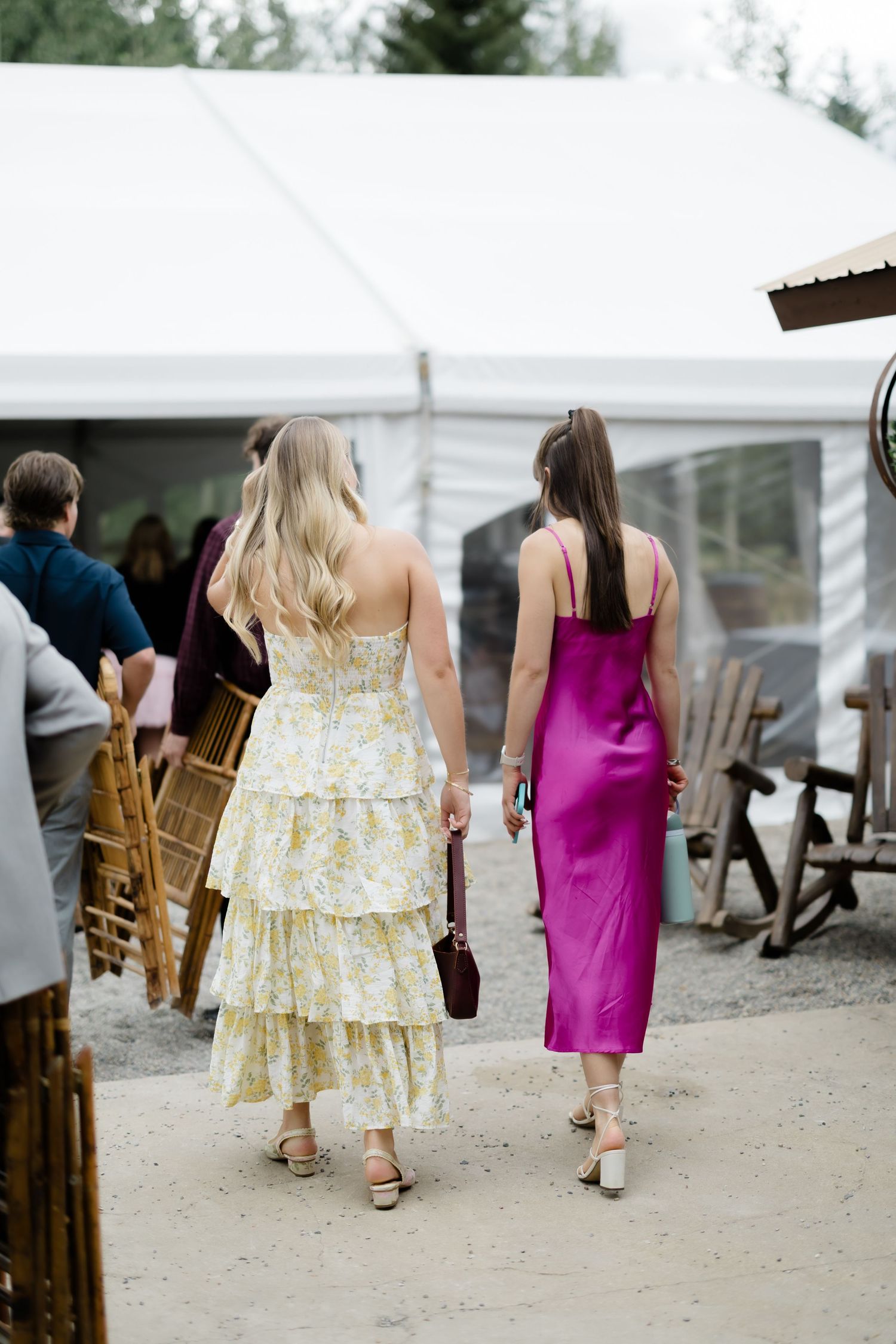 Two women in elegant dresses walk toward a wedding tent, one in floral and one in magenta satin.