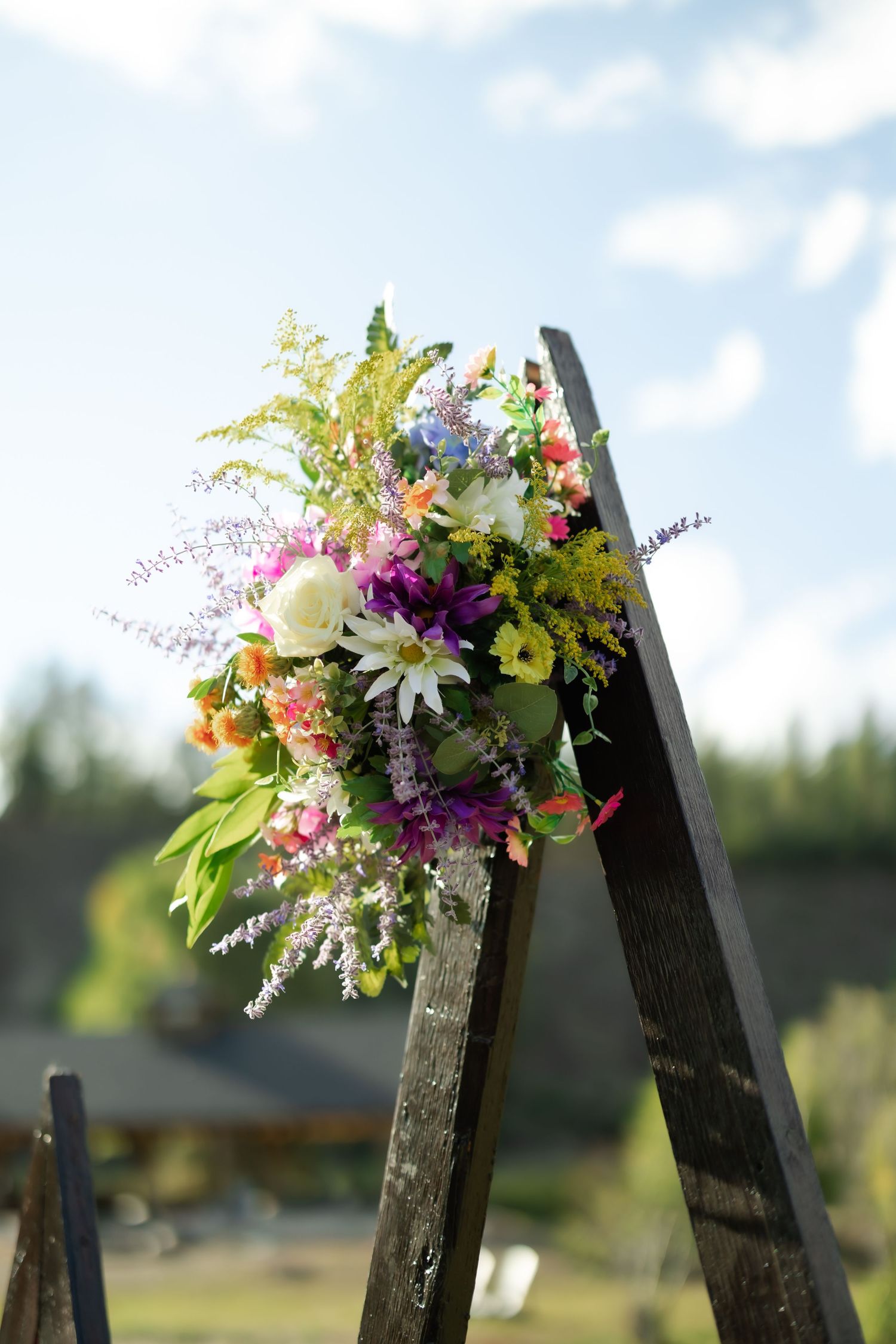 Colorful wildflower bouquet on rustic wooden ladder arch outdoors under blue sky for wedding ceremony.