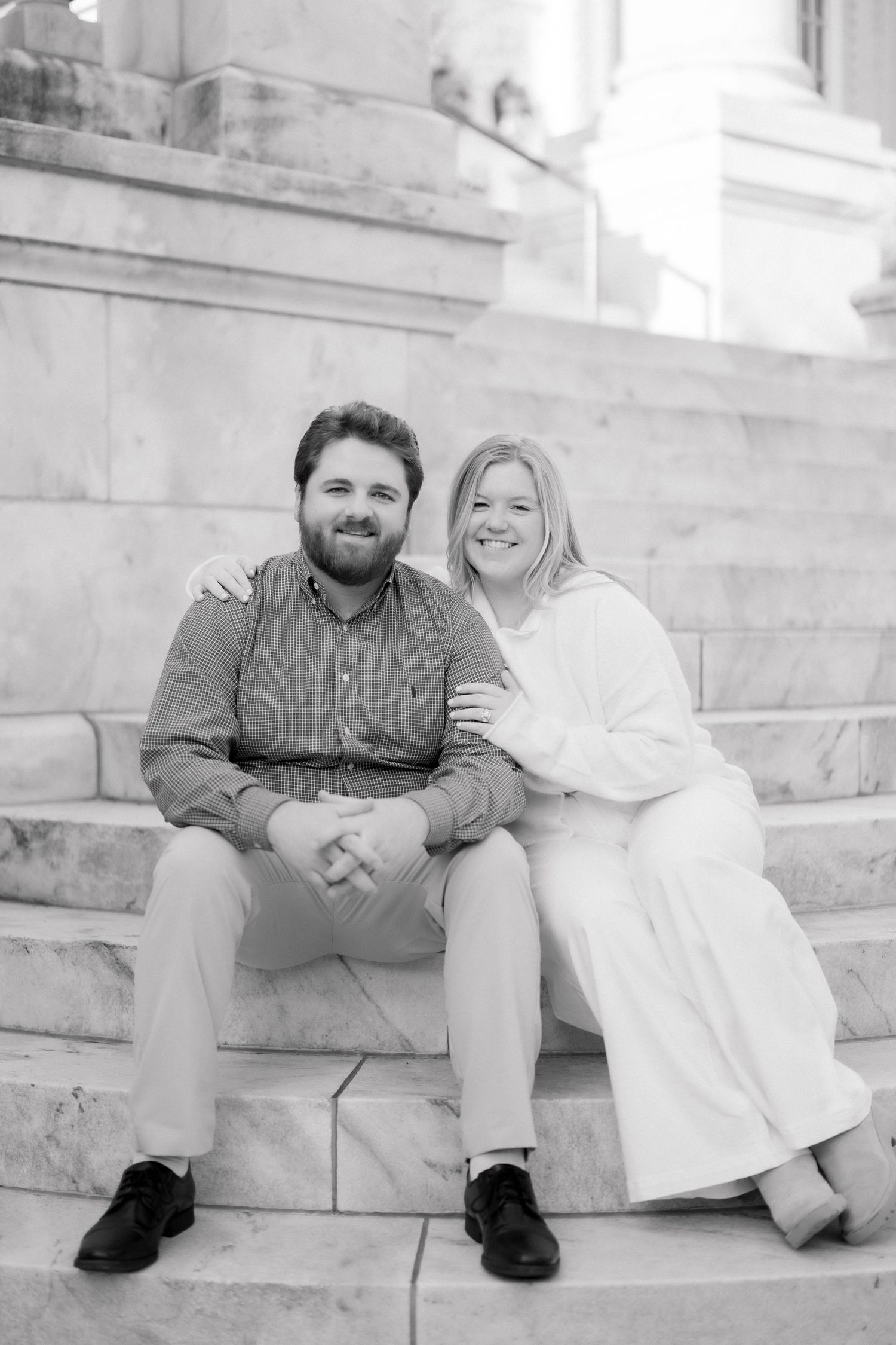 Black and white photo of happy engaged couple sitting on marble steps, smiling at camera.