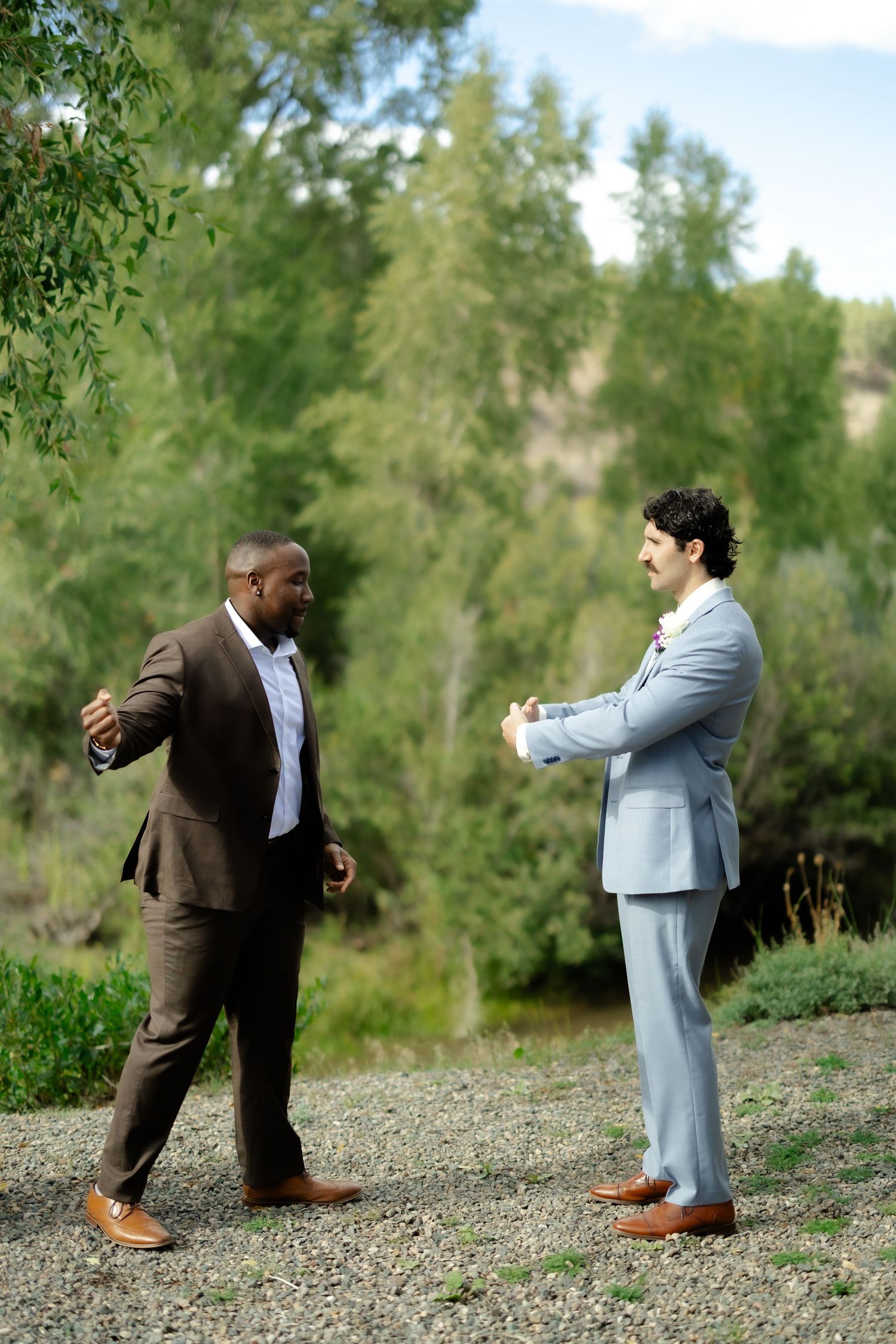 Two businessmen in suits conversing outdoors on gravel path surrounded by lush green trees.