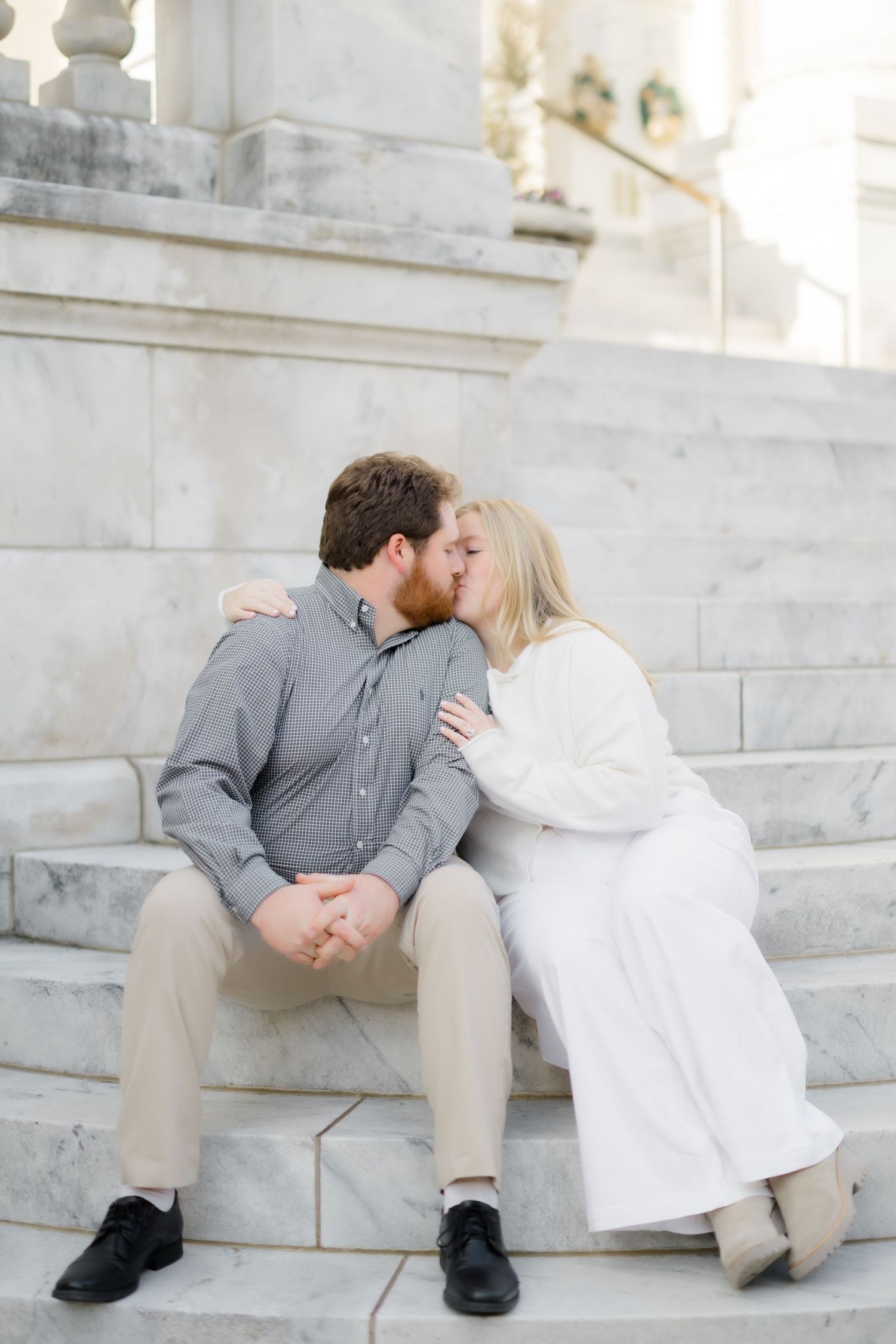 A couple shares a romantic kiss while sitting on marble steps during an outdoor engagement photo session.