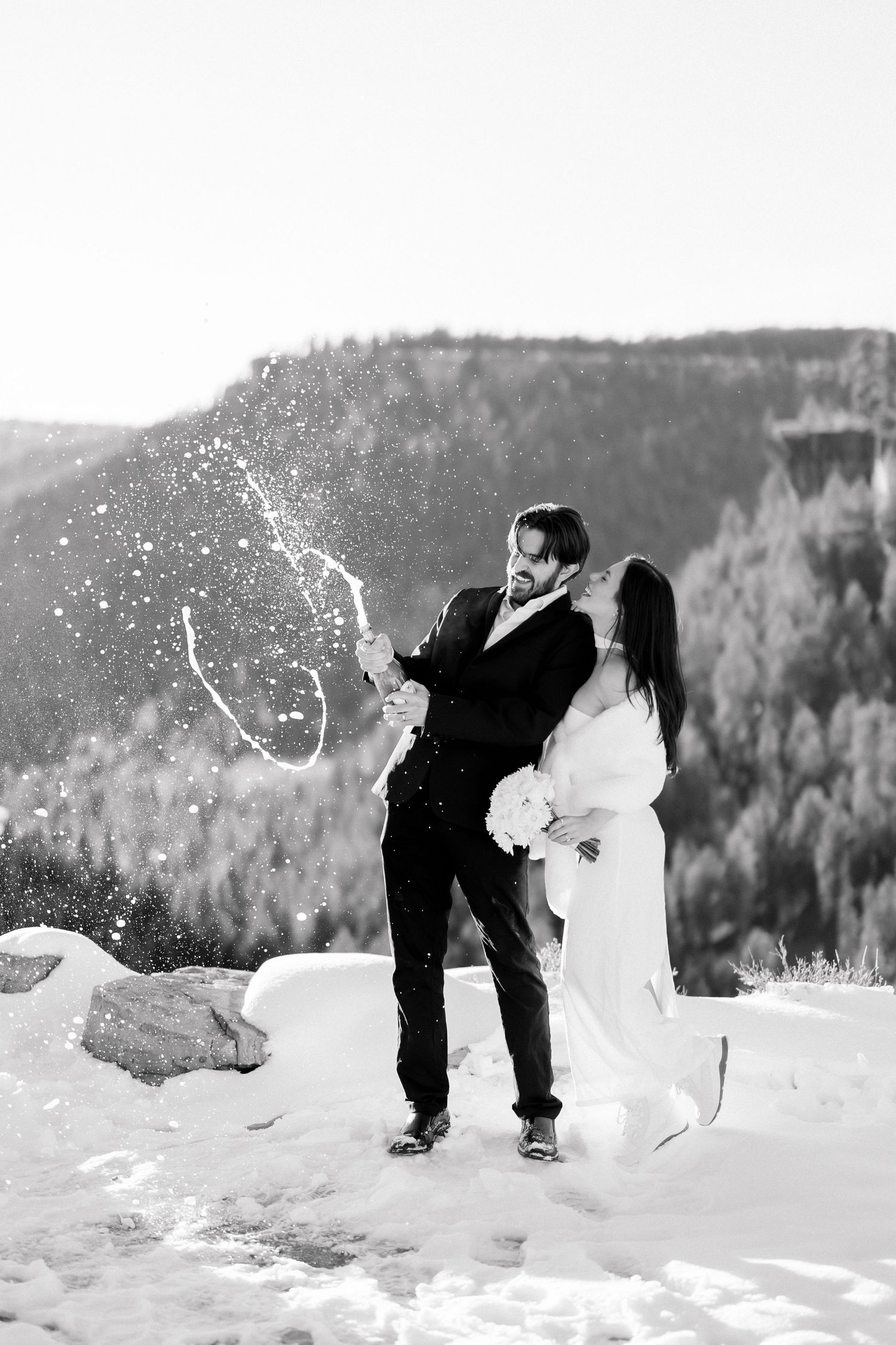 Black and white image of bride and groom popping champagne with snowy mountain forest backdrop.