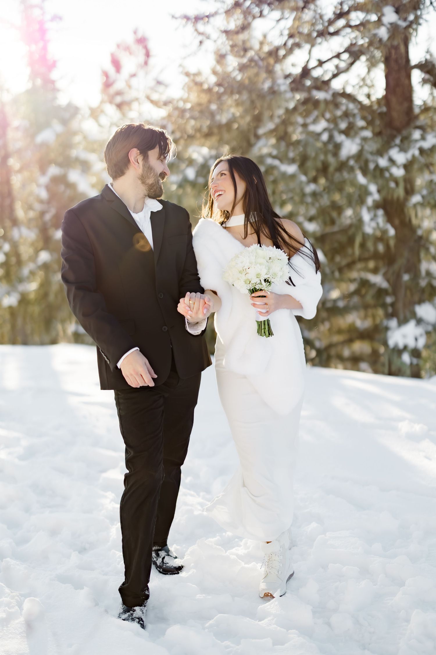Happy bride and groom walk hand in hand through snow-covered forest in winter wedding attire.
