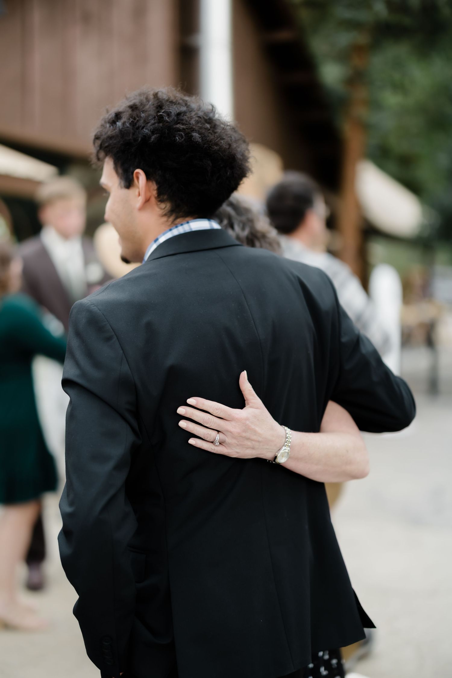 A guest places their hand on the back of a man in a black suit jacket at an outdoor wedding reception.