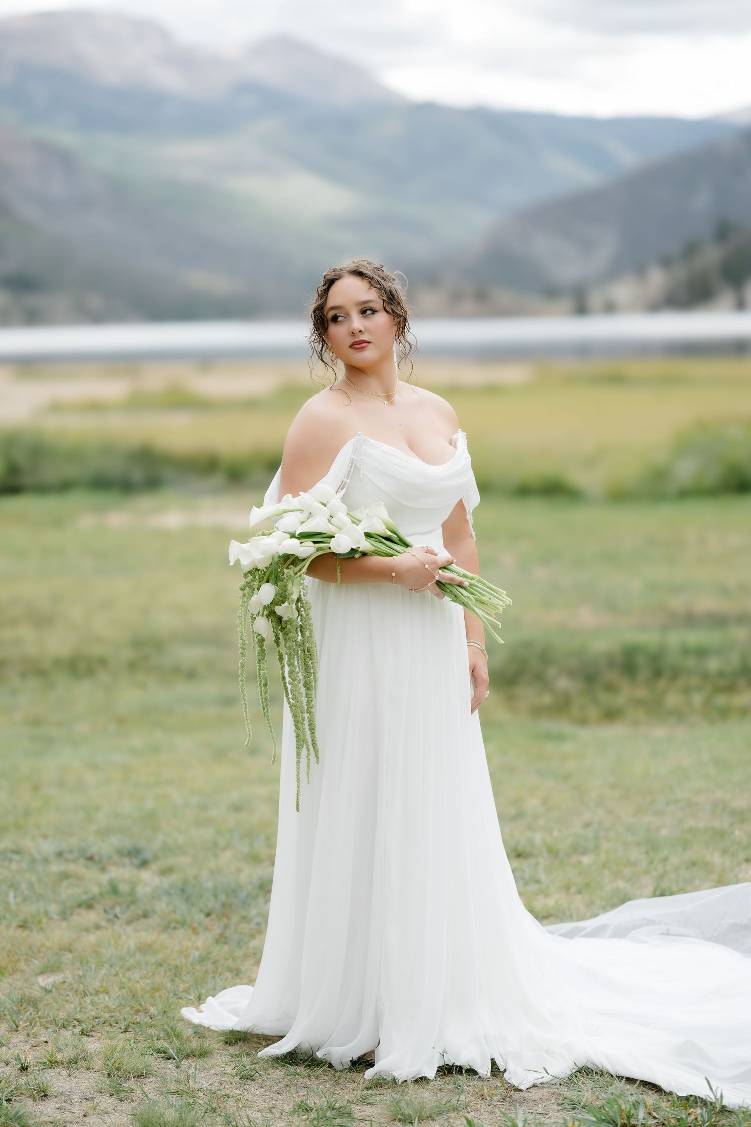 Bride in off-shoulder white gown holds calla lily bouquet in mountain meadow setting.