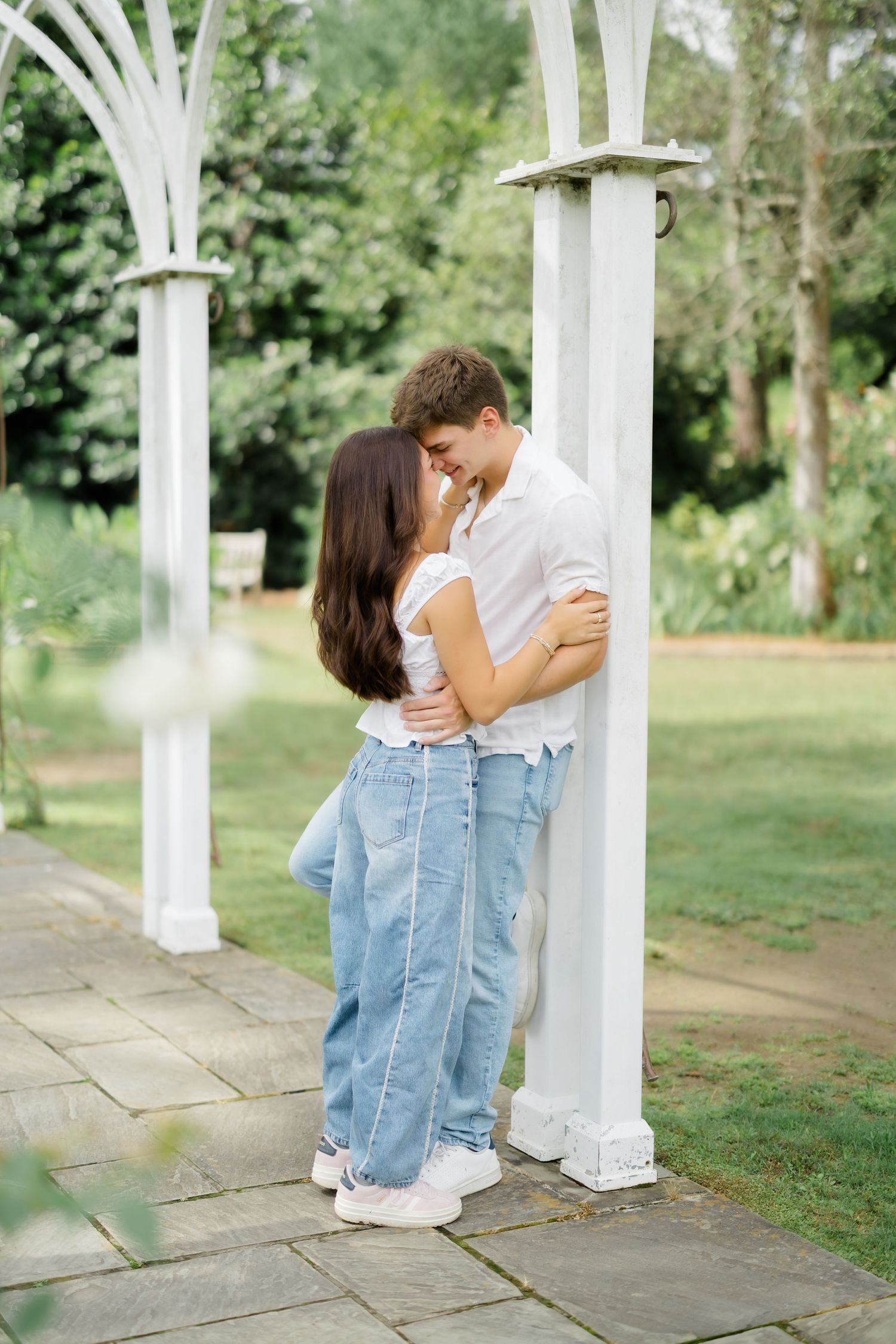 A young couple sharing a romantic moment leaning against a white garden arbor surrounded by lush greenery.