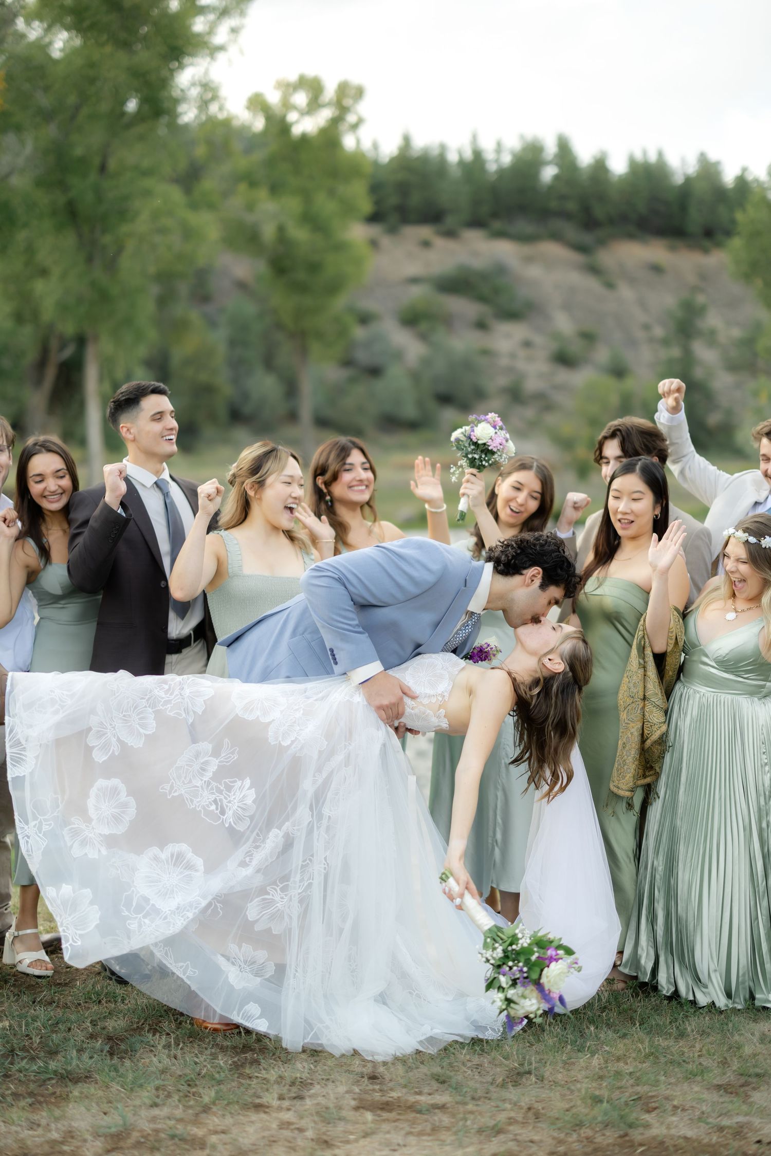 Groom dips bride for a kiss surrounded by cheering bridesmaids in sage green dresses and groomsmen outdoors.