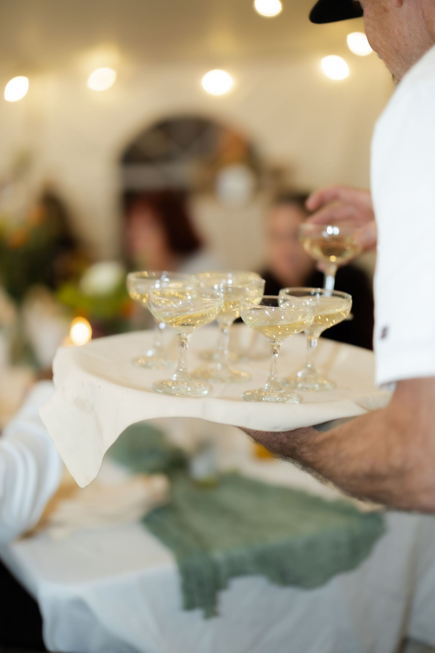 Waiter carrying a tray of champagne coupe glasses at an elegant event, with string lights in the background.