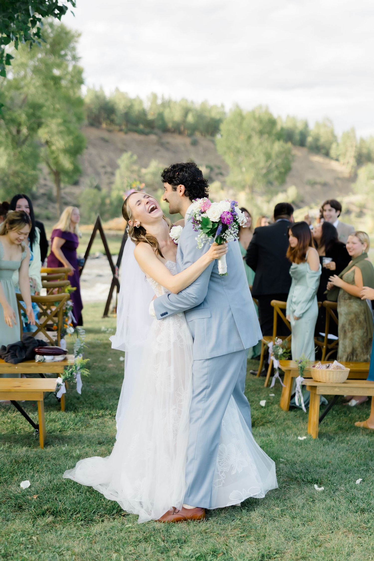 Joyful bride and groom embracing and laughing after outdoor wedding ceremony with guests seated on wooden chairs.